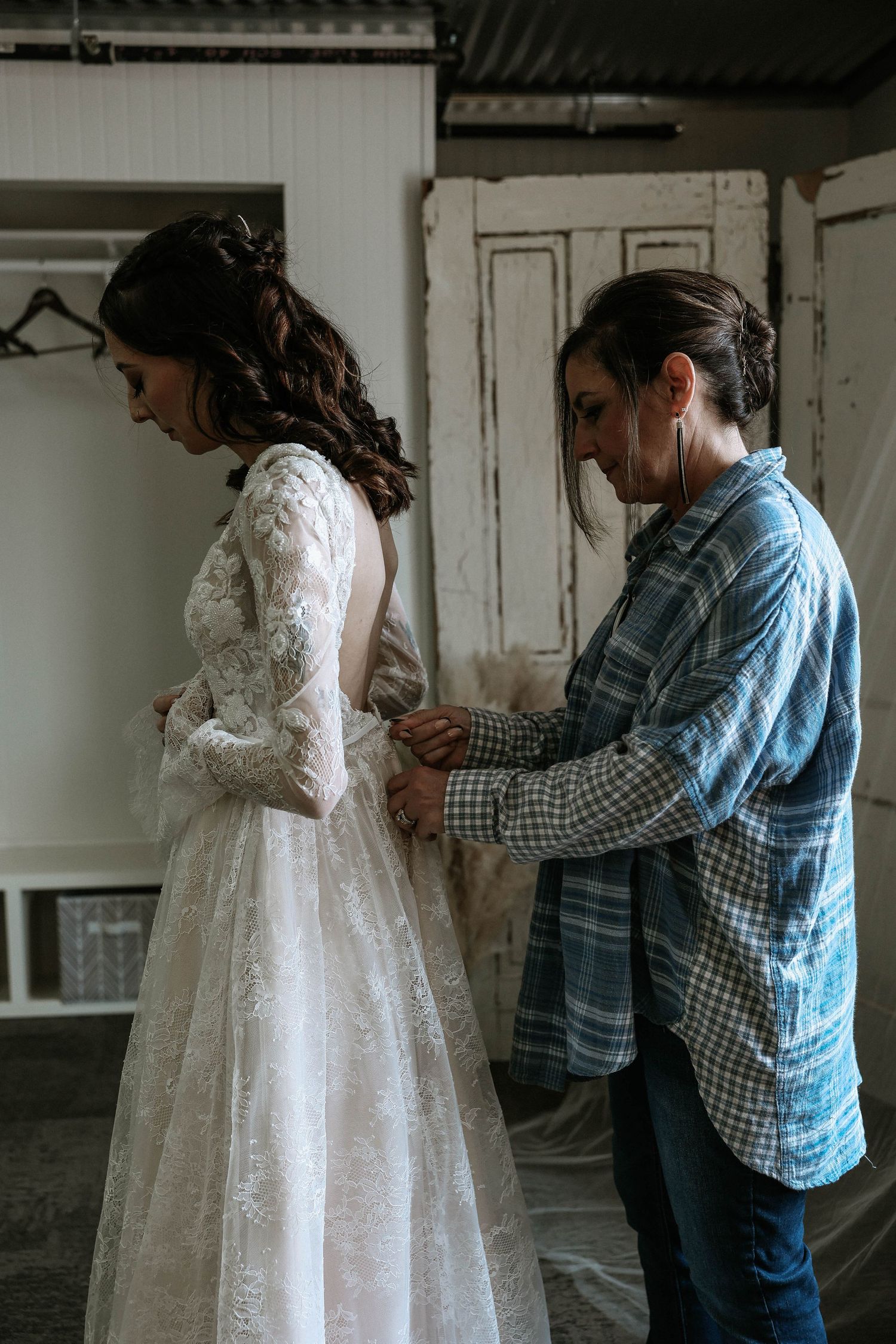 A woman in a plaid shirt helps a bride button up her lace wedding dress in a rustic room.