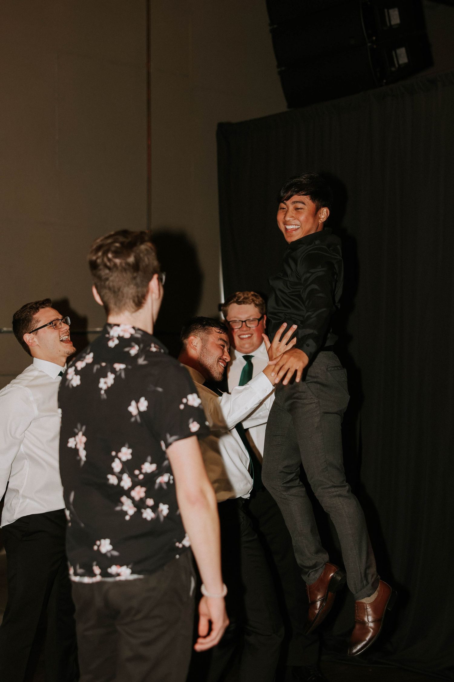 Group of young men in formal attire celebrating, lifting a smiling friend at a party or wedding reception.