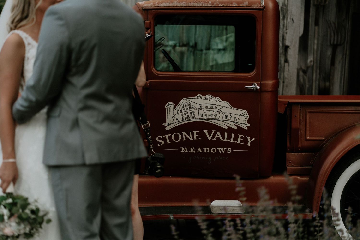 Bride and groom stand beside a vintage red Stone Valley Meadows truck at an outdoor wedding venue.