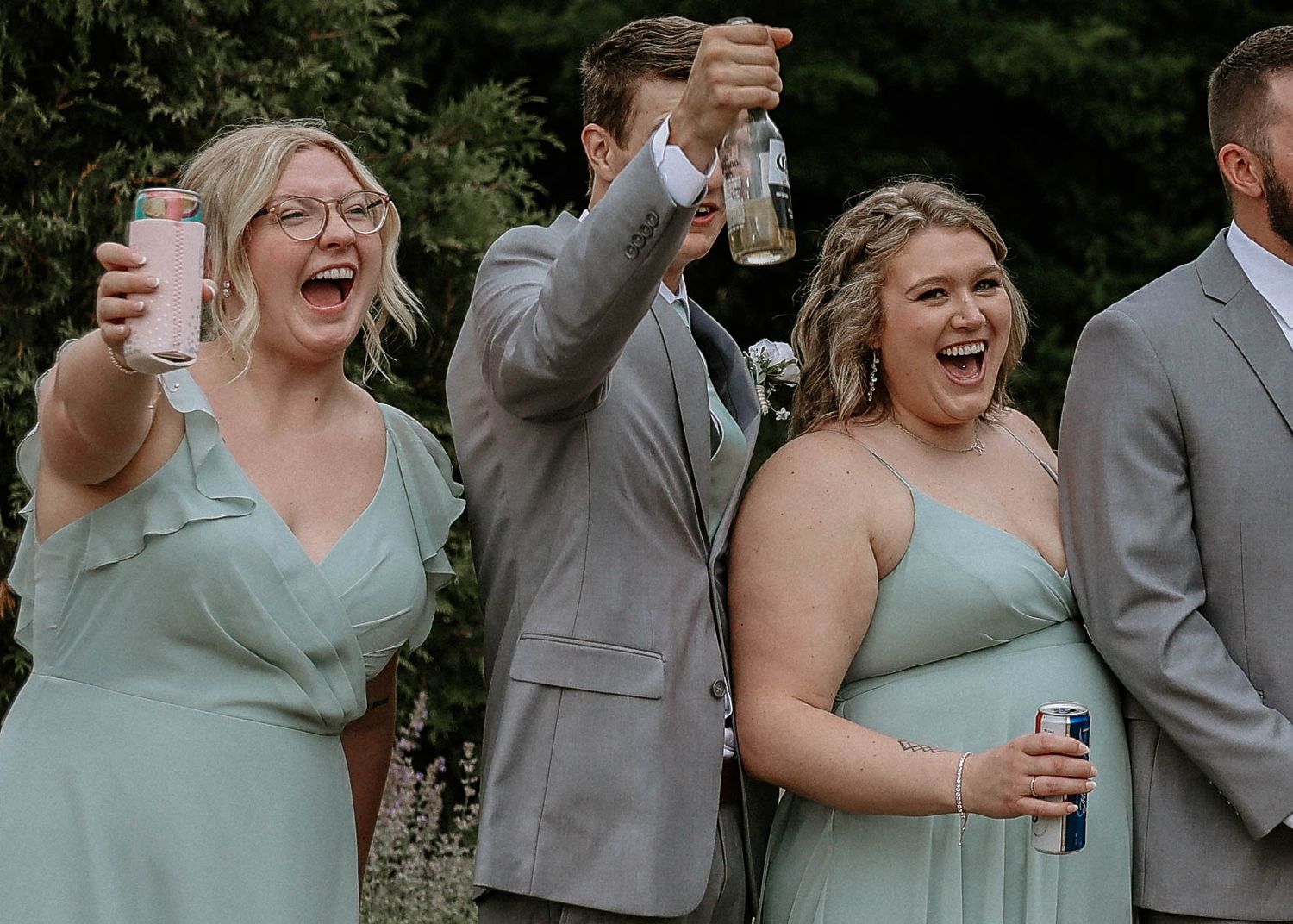 Two bridesmaids in sage green dresses raise drinks and laugh joyfully at an outdoor wedding celebration.