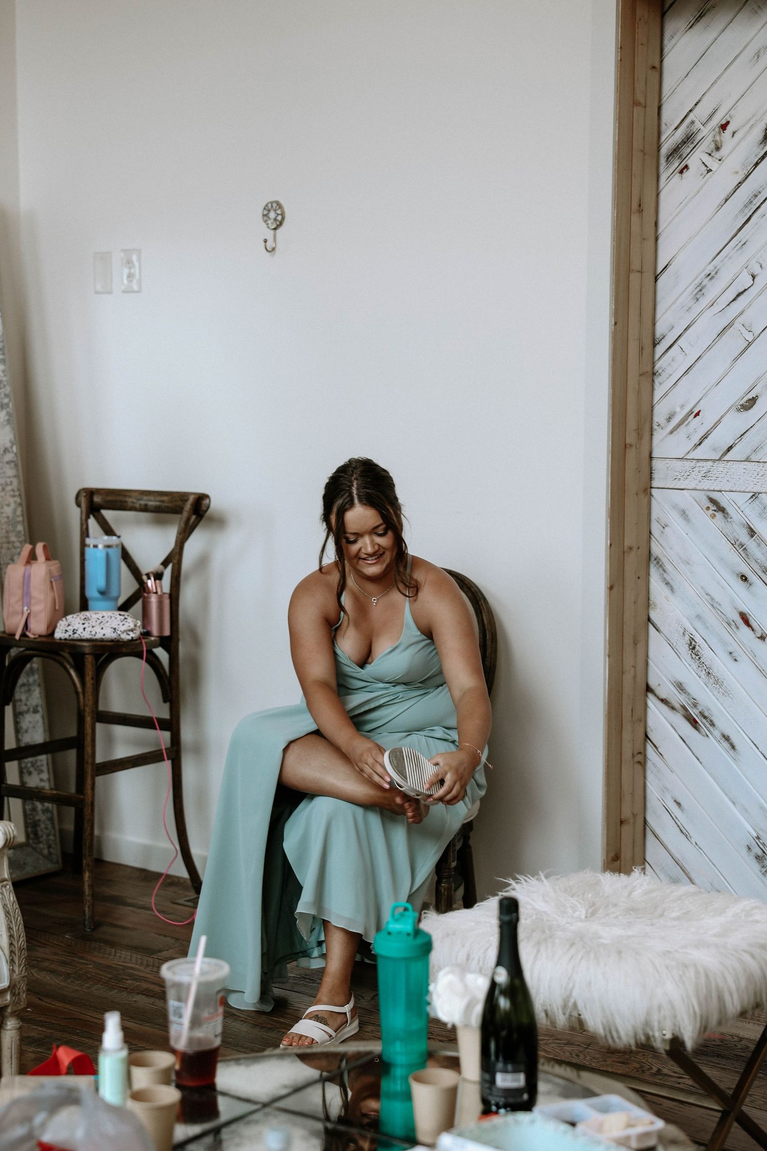 Bridesmaid in sage green dress putting on shoes in a rustic getting-ready room before a wedding.