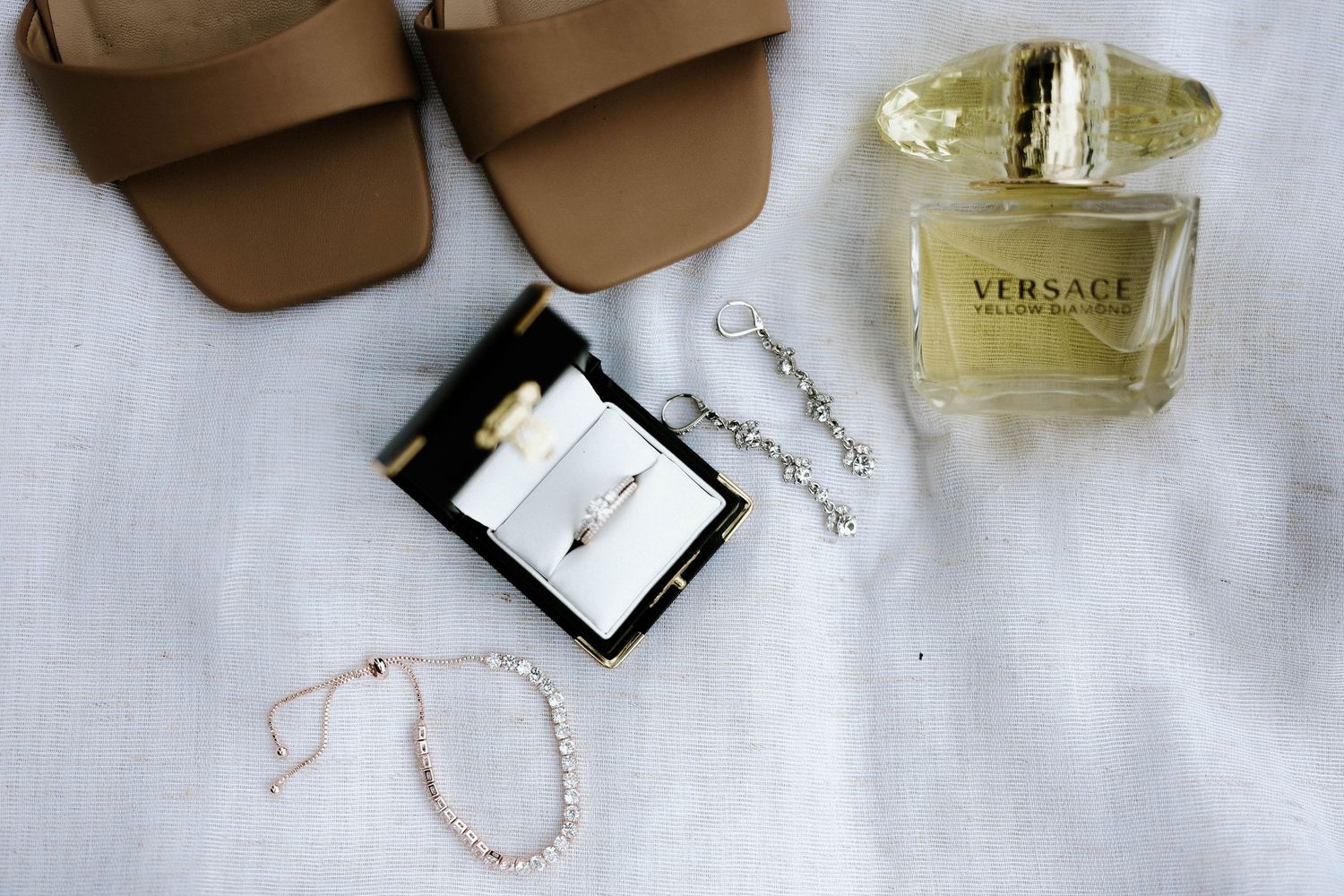 Flatlay of Versace Yellow Diamond perfume, diamond ring, pearl bracelet, and tan sandals on white fabric.