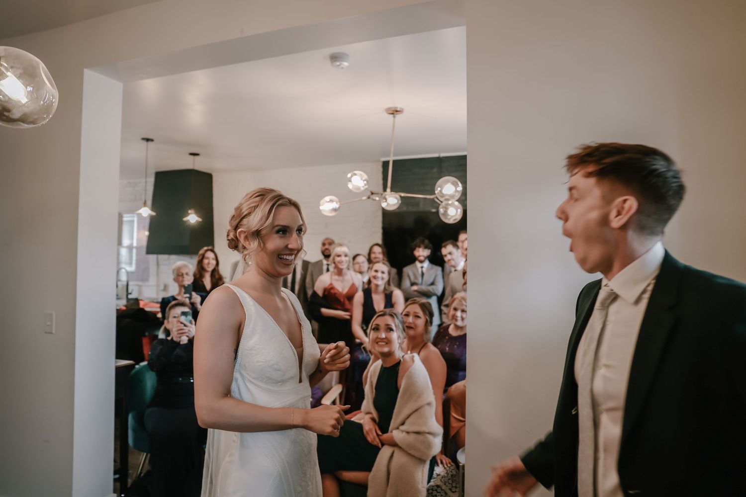 Bride and groom exchange vows indoors with wedding guests gathered in background during intimate ceremony.