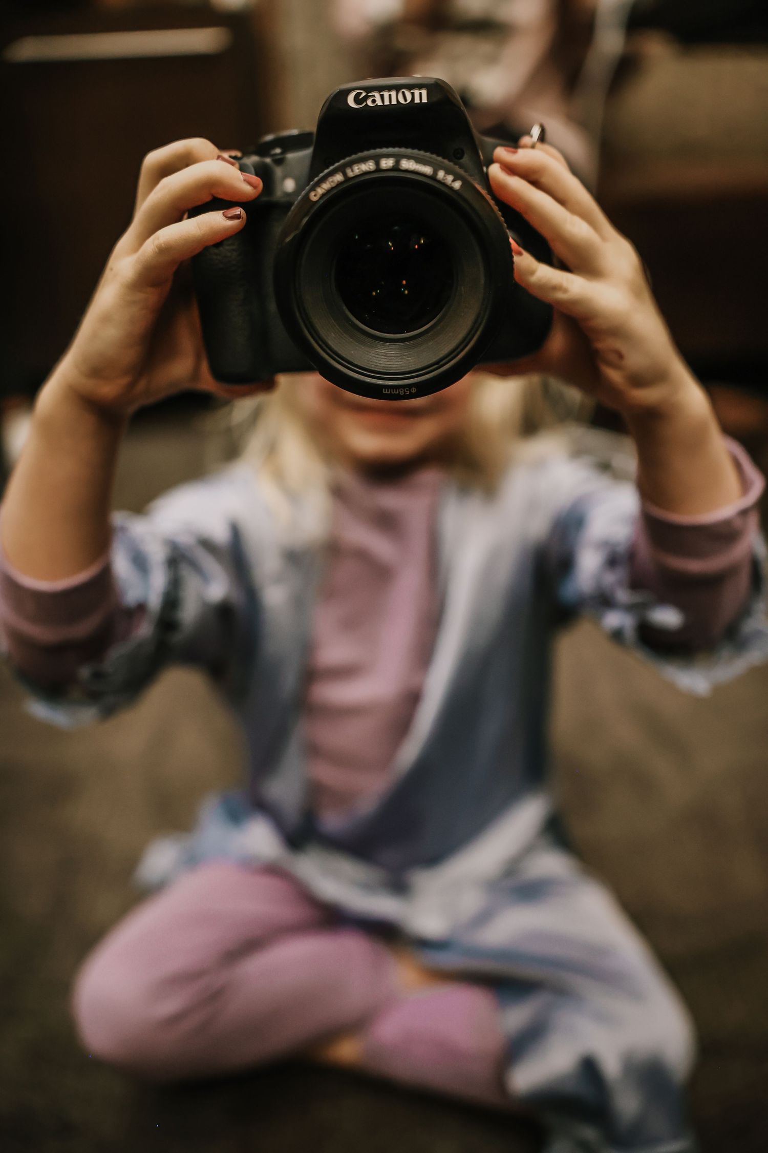 Young girl sitting cross-legged holding a Canon DSLR camera up to her face, obscuring her features.