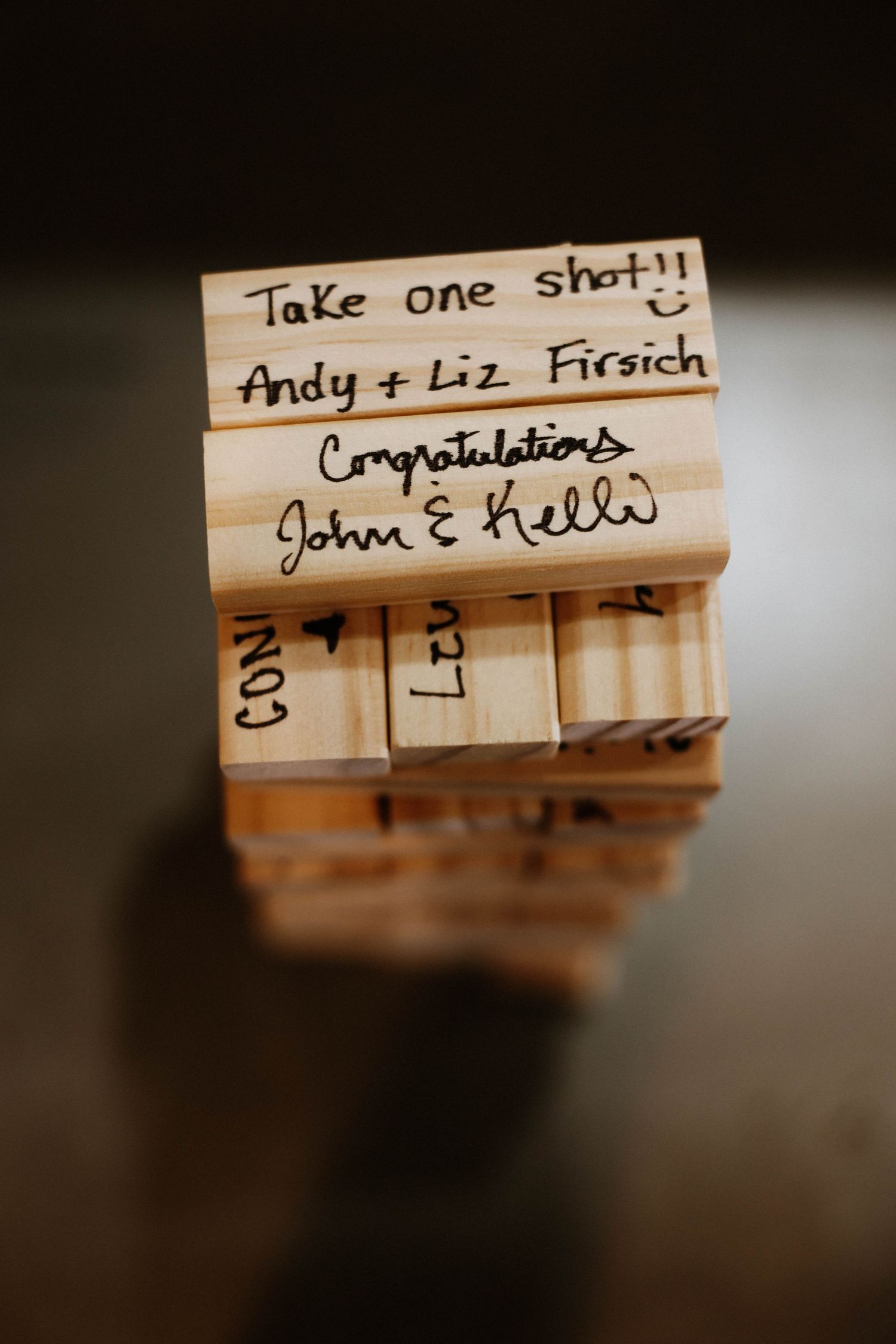 Jenga blocks inscribed with wedding messages including Take one shot, congratulations, and names Andy, Liz, John, and Kelli.