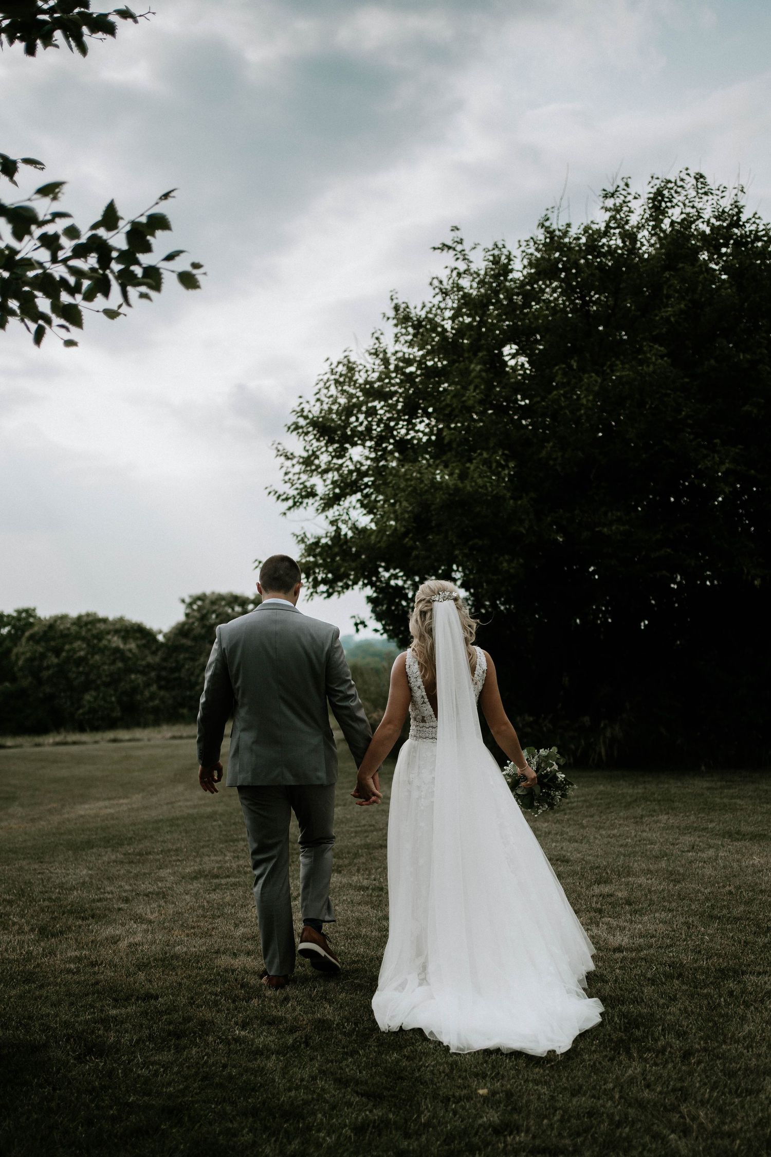 Bride and groom walking hand in hand across green field toward trees under overcast sky.