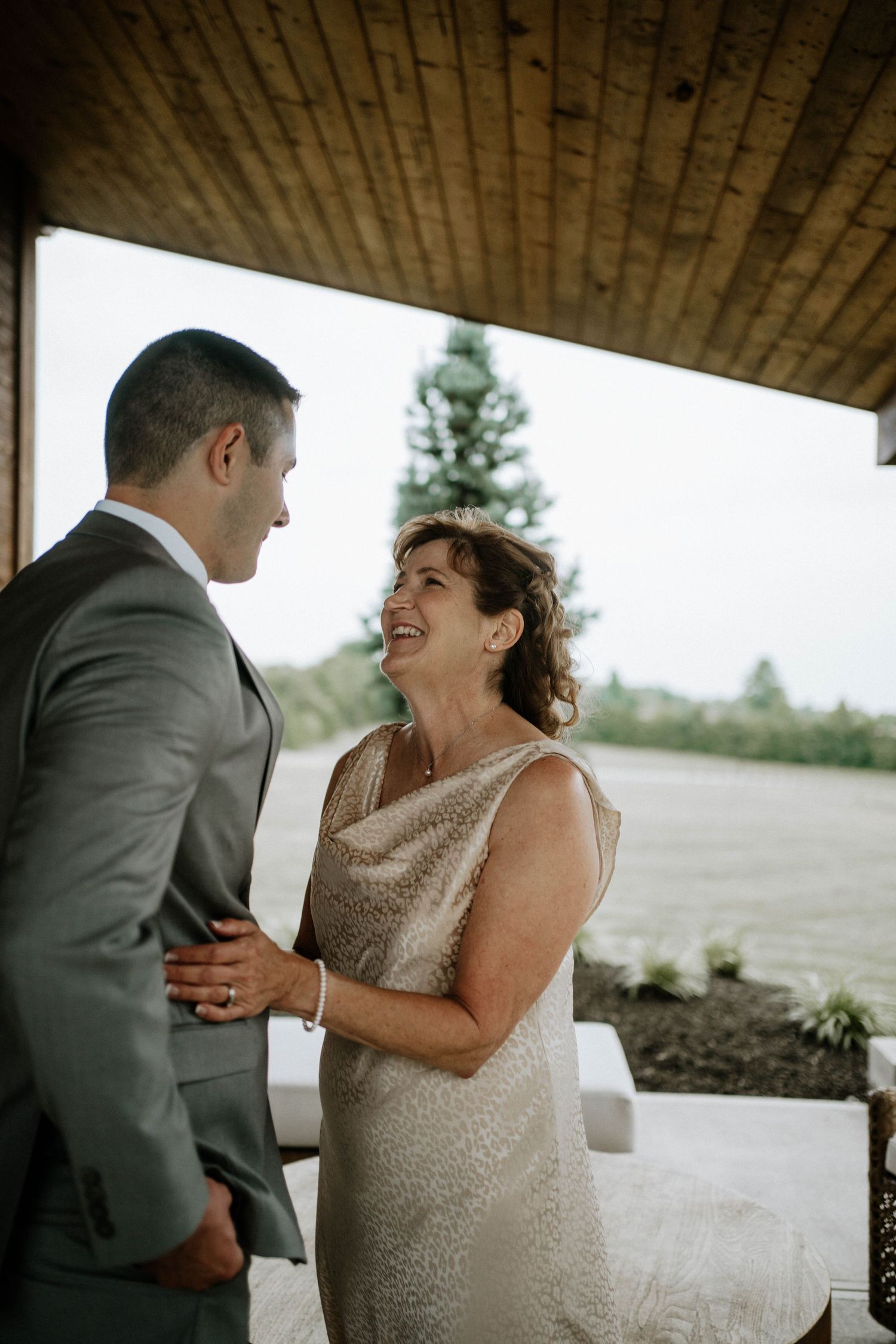 Groom in gray suit sharing joyful moment with smiling older woman in gold dress.