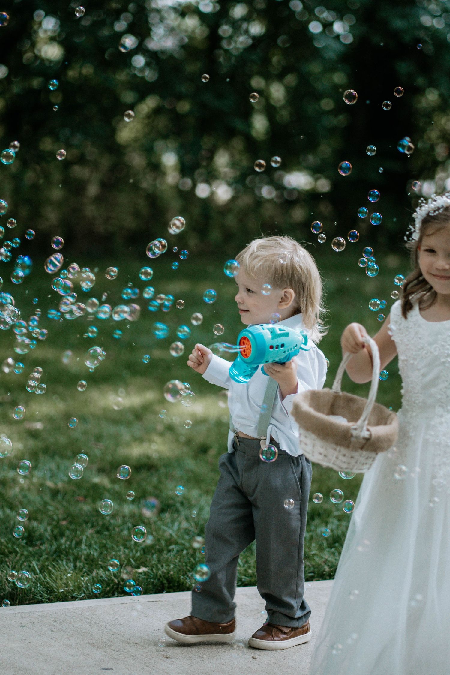 Young boy using a bubble gun at a wedding while a flower girl in white dress holds a basket outdoors.