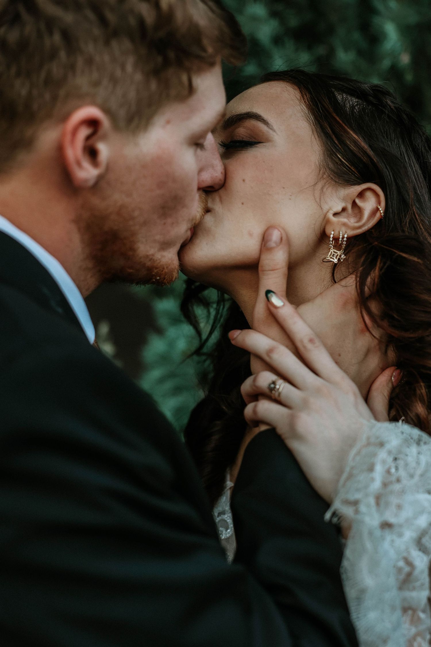 A couple shares a romantic kiss, the bride wearing lace and gold earrings, groom in dark suit, in a moody outdoor wedding portrait.