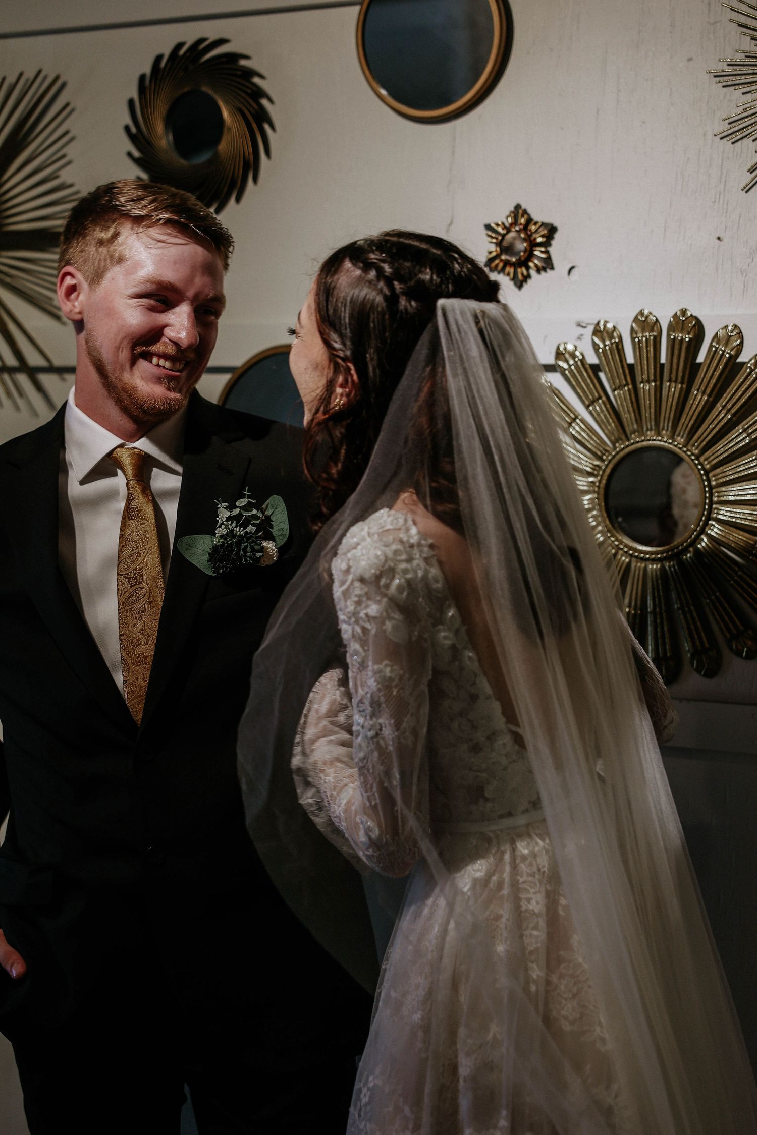 Groom in black suit with gold tie smiles at bride in lace wedding dress and veil, surrounded by decorative gold mirrors.