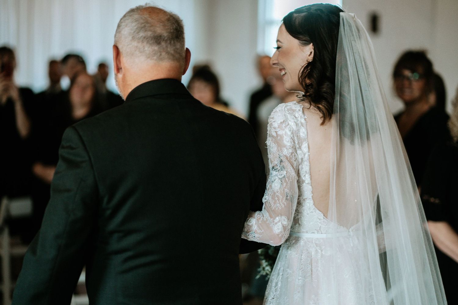 Bride in lace gown with veil walks down aisle with older man in black suit during wedding ceremony.