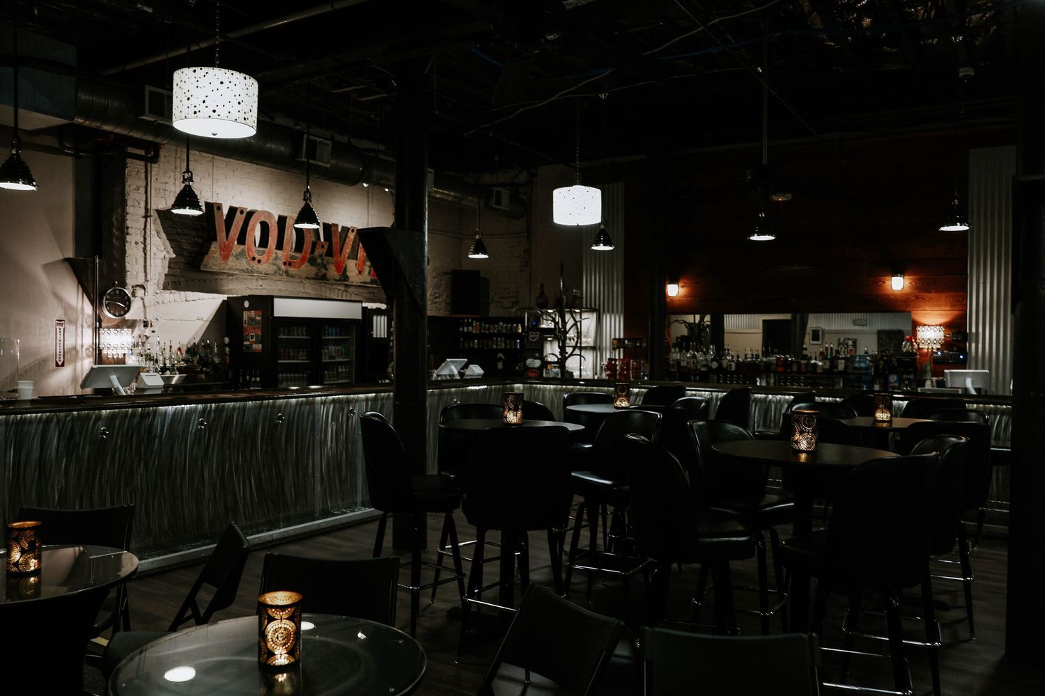 Dimly lit upscale bar and restaurant interior with dark furniture, pendant lights, and a glowing red neon sign on the wall.
