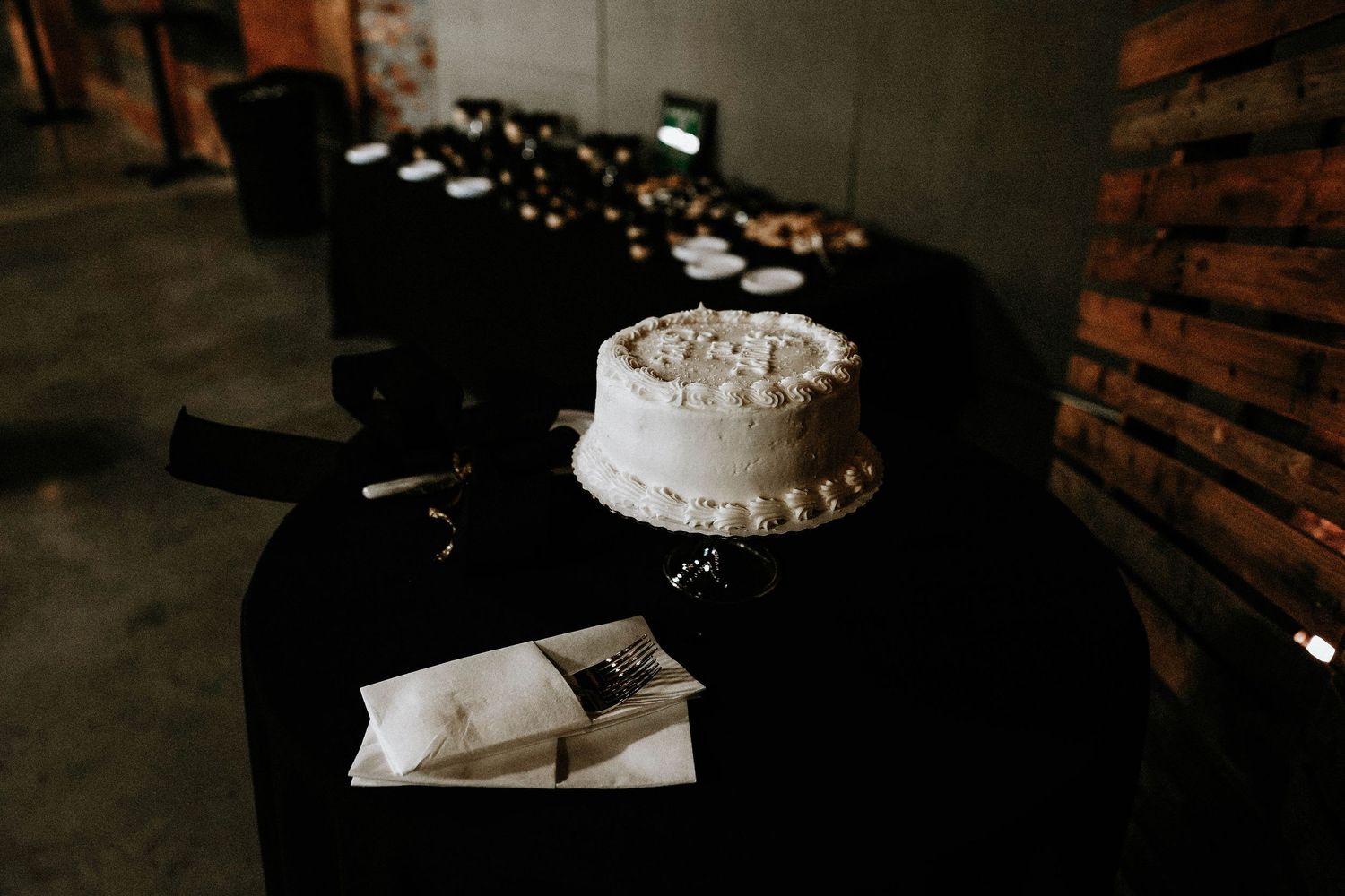 White frosted wedding cake on black draped table in dimly lit rustic venue with bokeh lights in background.