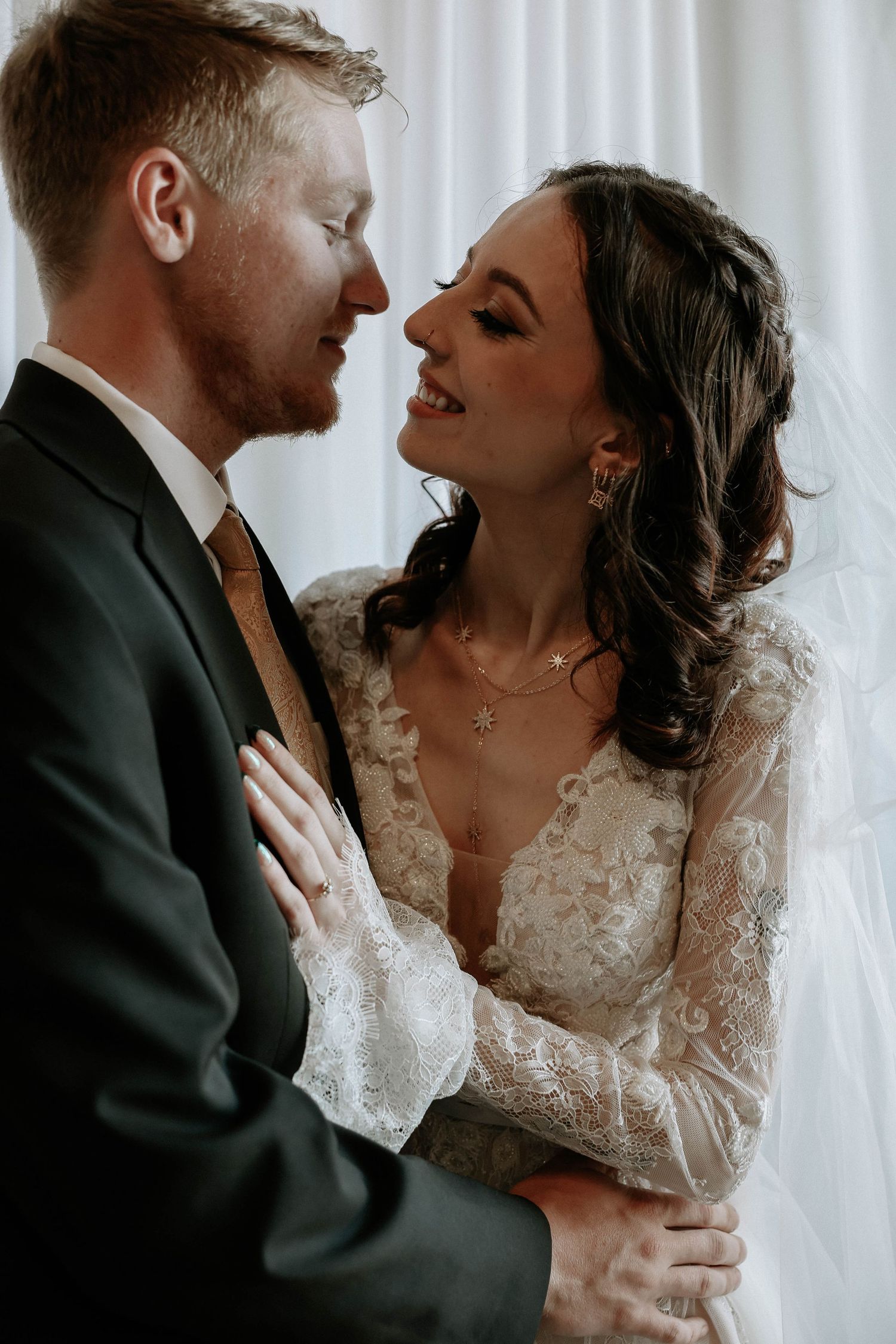 A joyful bride in lace gown embraces her groom in a suit, sharing a romantic moment on their wedding day.