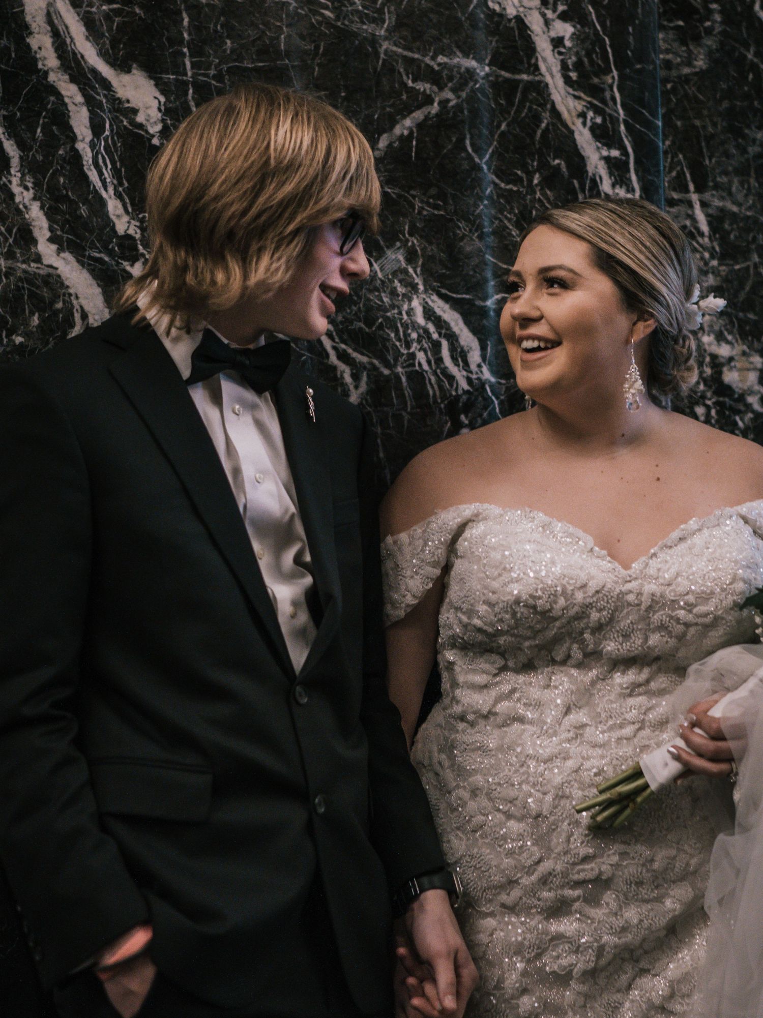 Newlyweds laugh together in front of marble wall, bride in beaded gown and groom in black tuxedo.