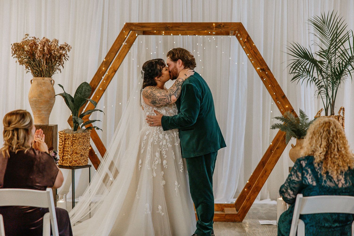 Bride and groom share first kiss under wooden hexagon arch with fairy lights at indoor wedding ceremony.