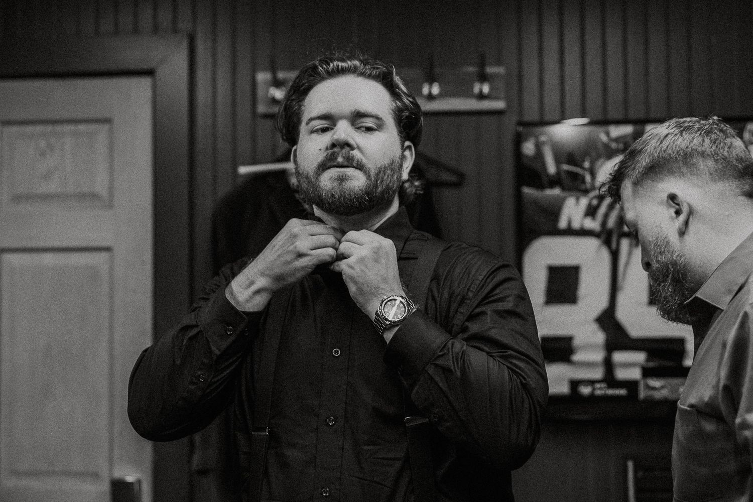 Black and white photo of bearded groom adjusting bow tie with groomsman assisting nearby.