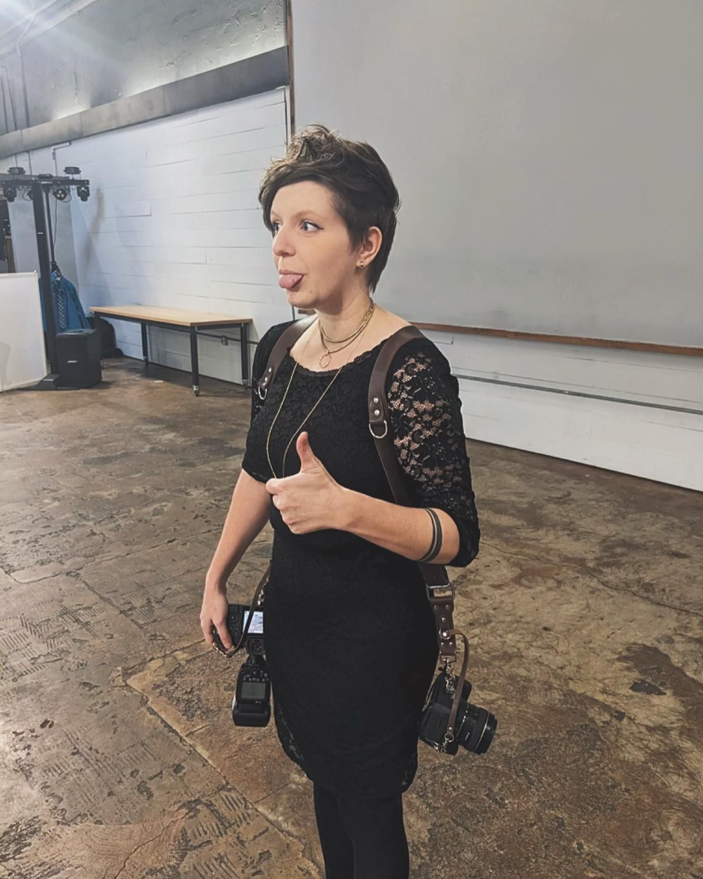 Female wedding photographer in black outfit giving thumbs up while holding camera at venue.