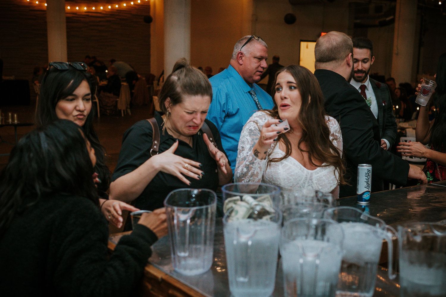 Bride interacting with guests at wedding reception bar with drinks and glasses on counter.