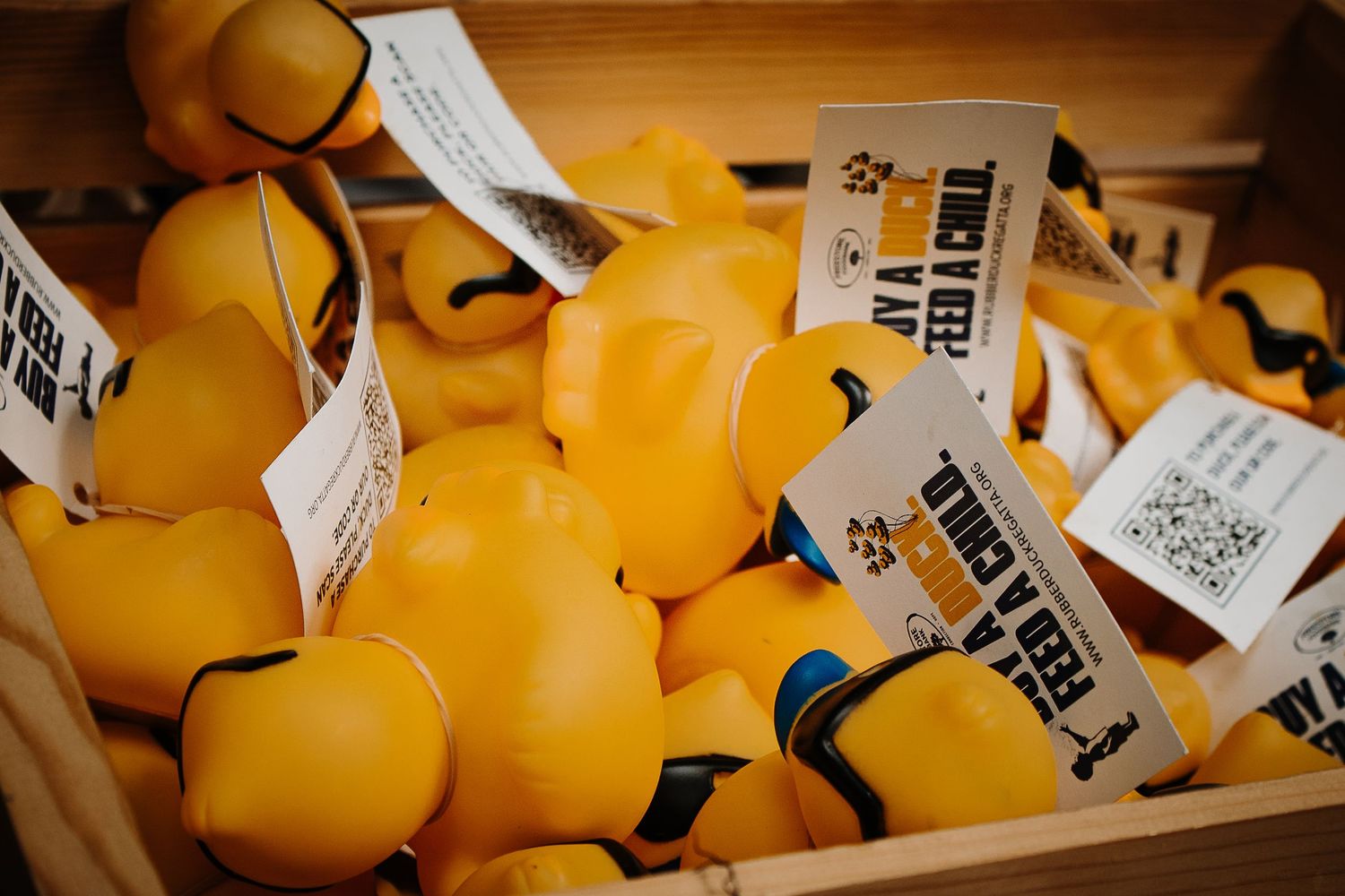 A wooden crate filled with yellow rubber ducks with attached tags reading 'Touch & Feel a Duck' at an event.
