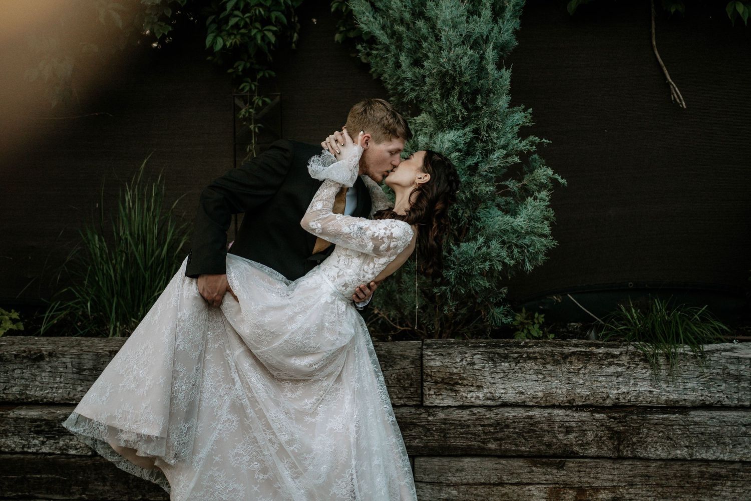 Bride and groom sharing a romantic dip kiss in front of evergreen trees, bride in lace gown.
