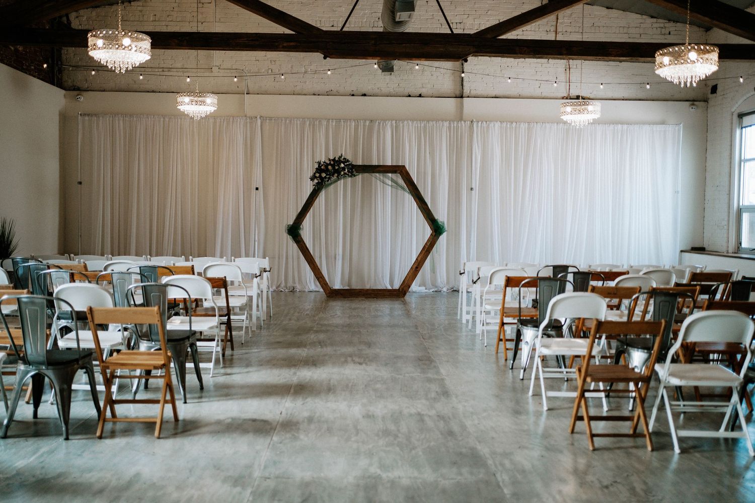 Elegant wedding ceremony setup with hexagonal arch, white draping, mixed chairs, and crystal chandeliers in a modern venue.