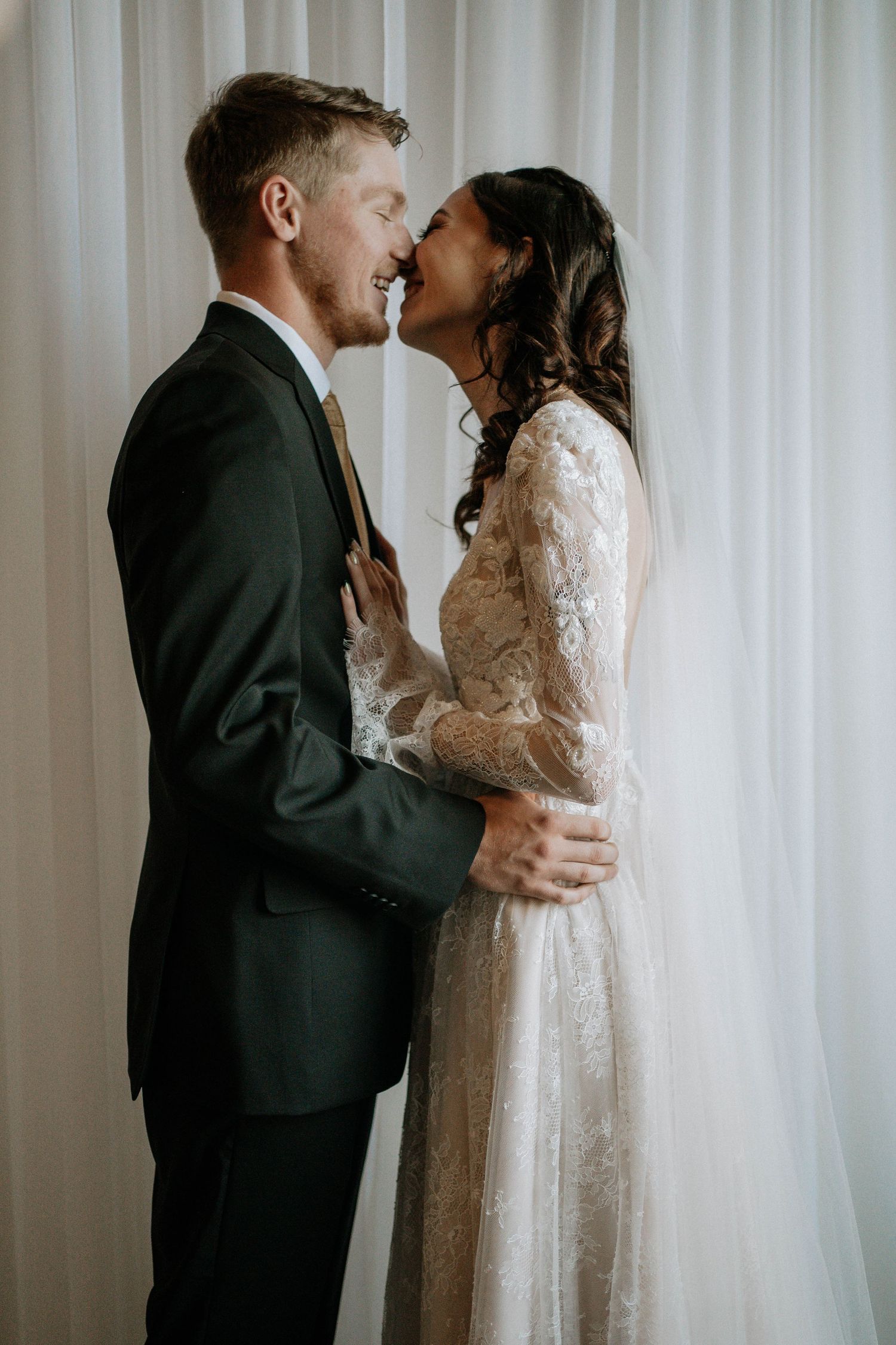 Joyful bride and groom sharing a romantic moment, laughing together in wedding attire before white curtains.