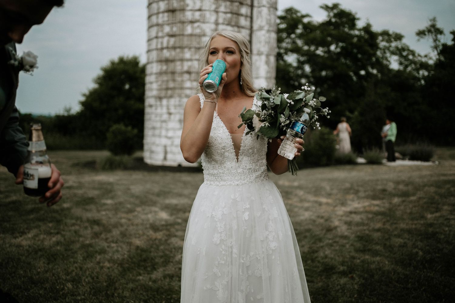 Blonde bride in lace gown drinking from cup while holding bouquet near rustic stone silo.