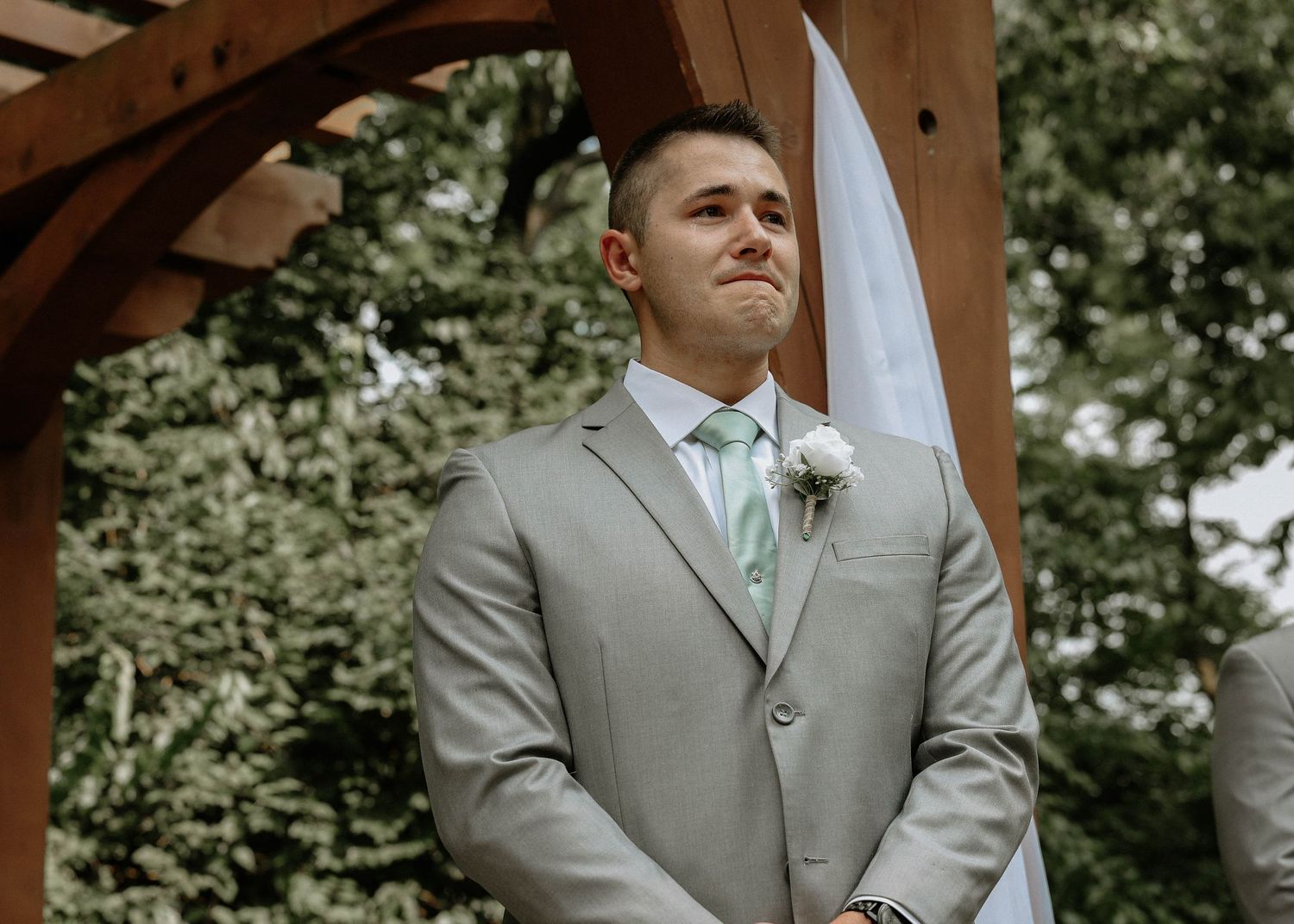 Emotional groom in gray suit with mint tie and boutonniere watching bride walk down the aisle outdoors.