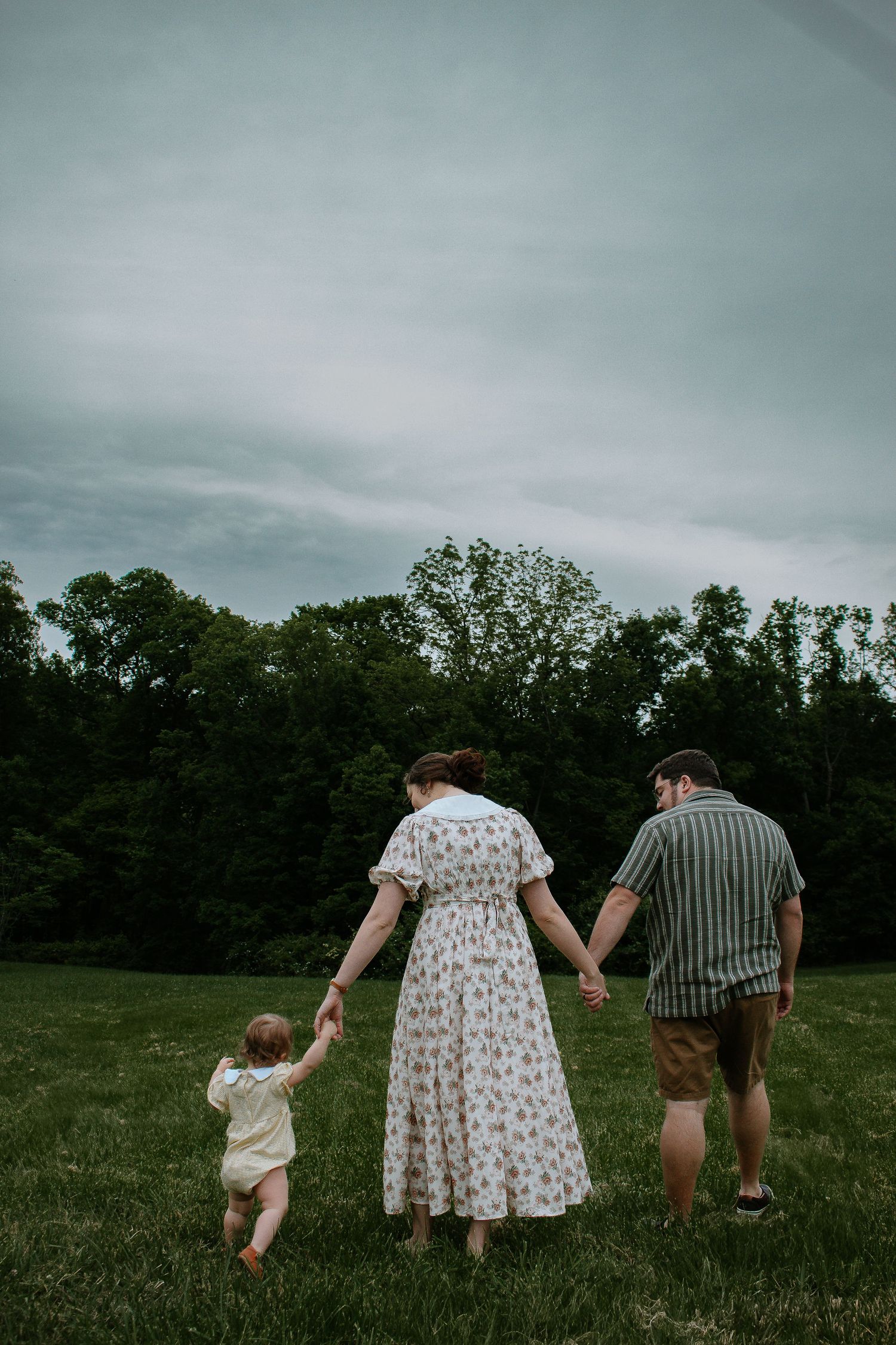 Young family walking hand-in-hand across green meadow with toddler, viewed from behind at French Park in Cincinnati, Ohio