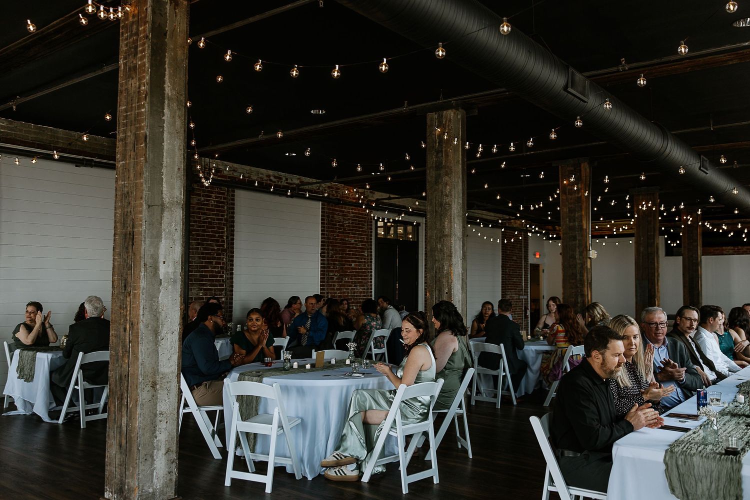 Guests seated at round tables with white linens under string lights in a rustic brick wedding reception venue.
