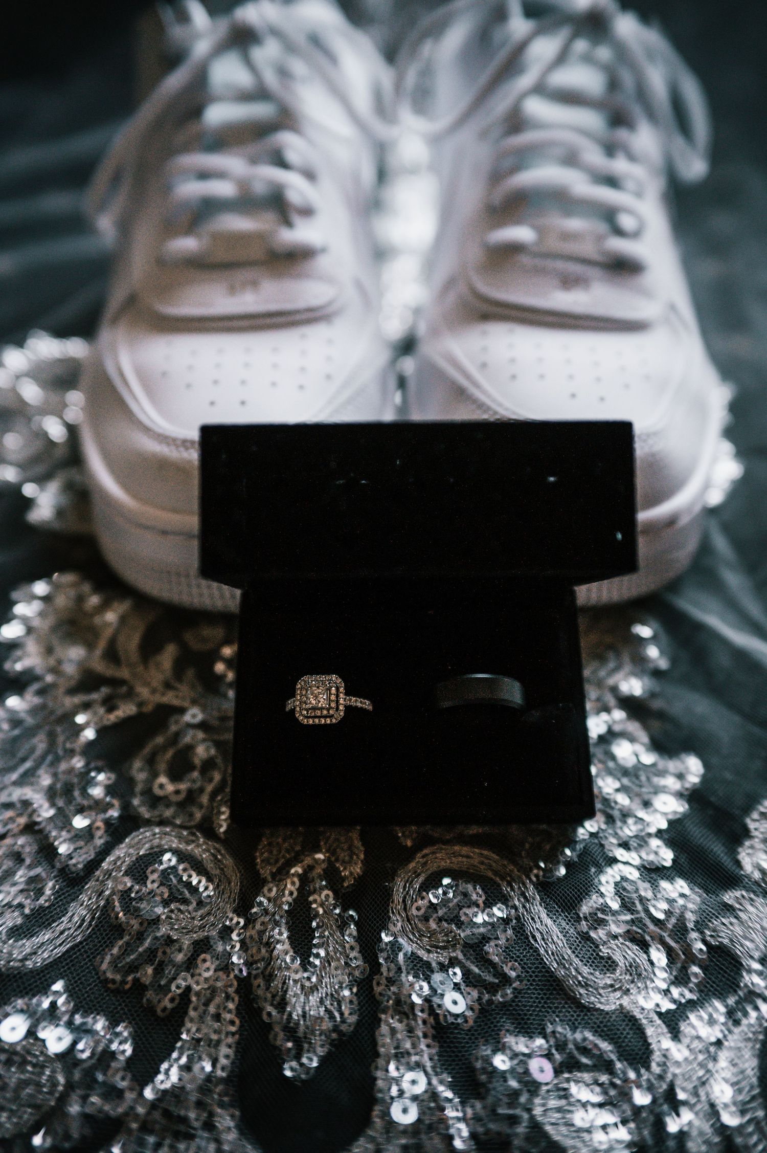 Wedding rings in black velvet box beside white Nike sneakers on sequined fabric.