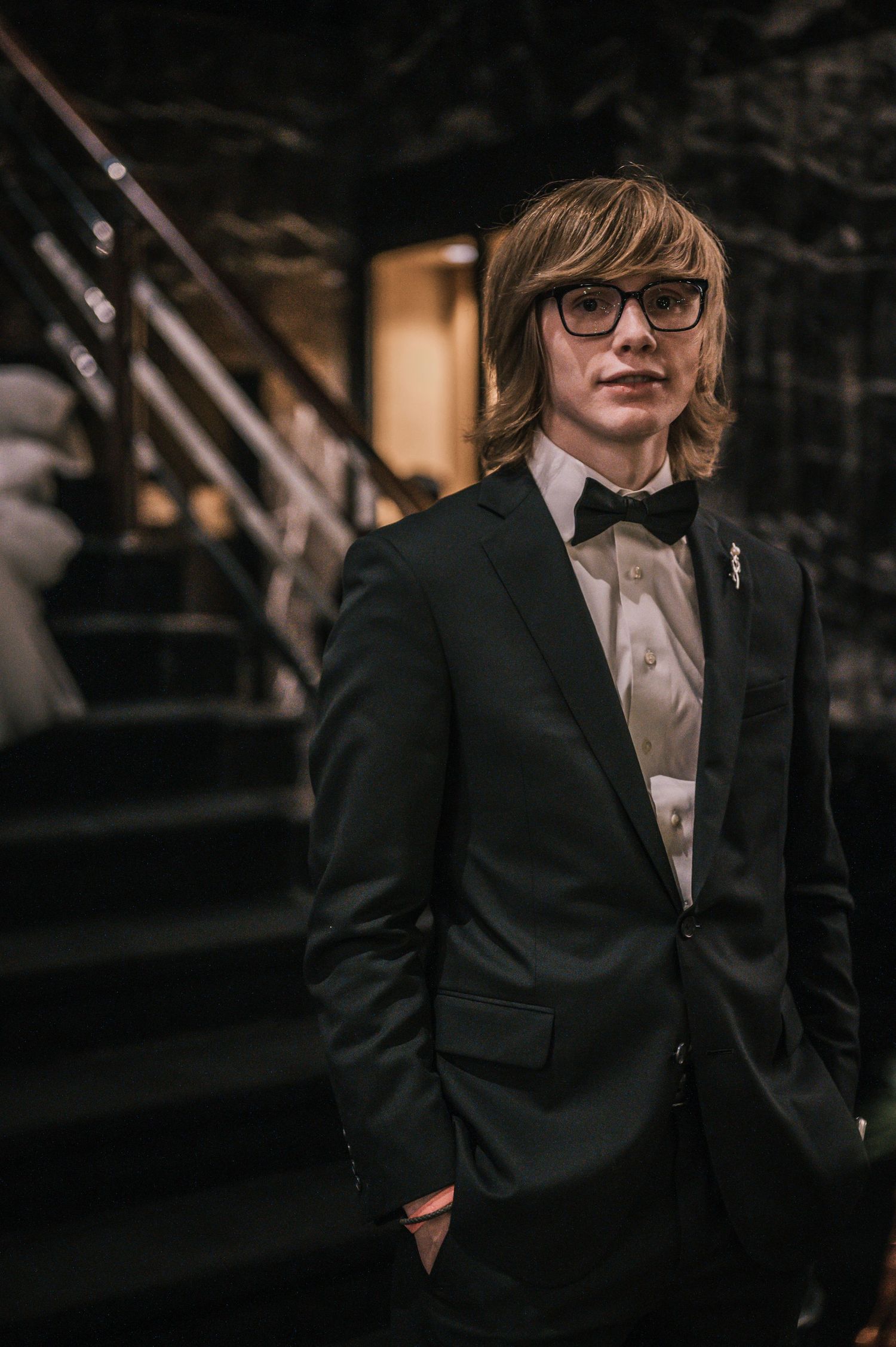 Young man in black tuxedo with bow tie and glasses poses on dark marble staircase.