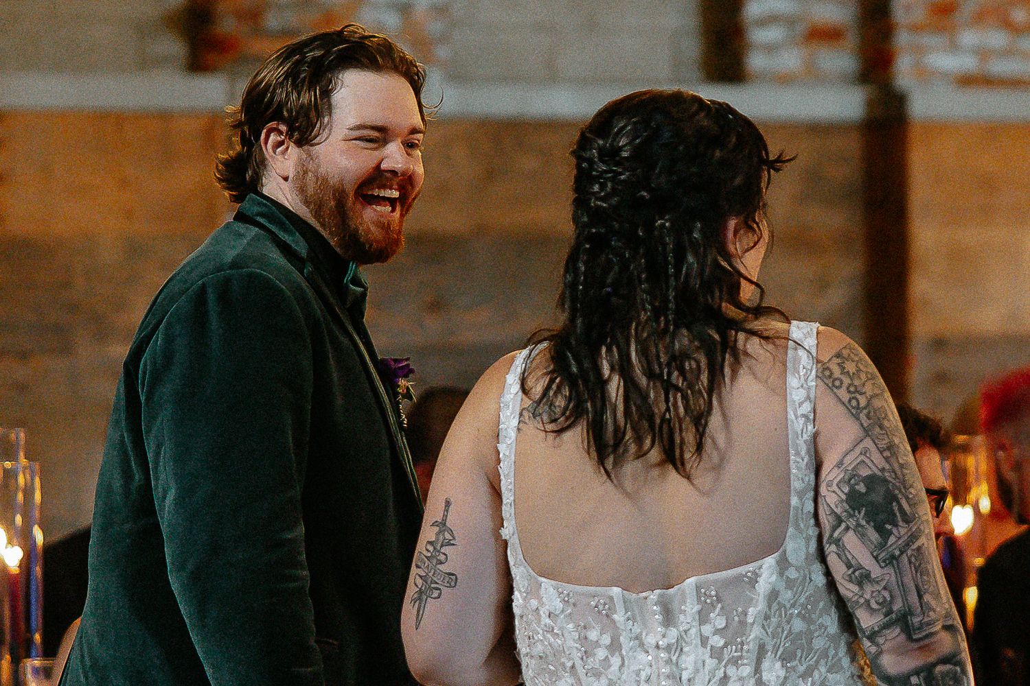 Smiling groom in green velvet suit laughing with tattooed bride at wedding reception.