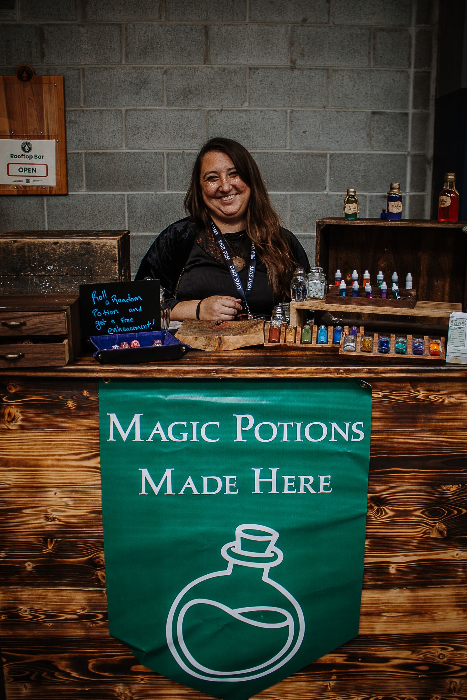 Smiling woman behind a wooden bar with a green Magic Potions Made Here banner at a fantasy-themed event.