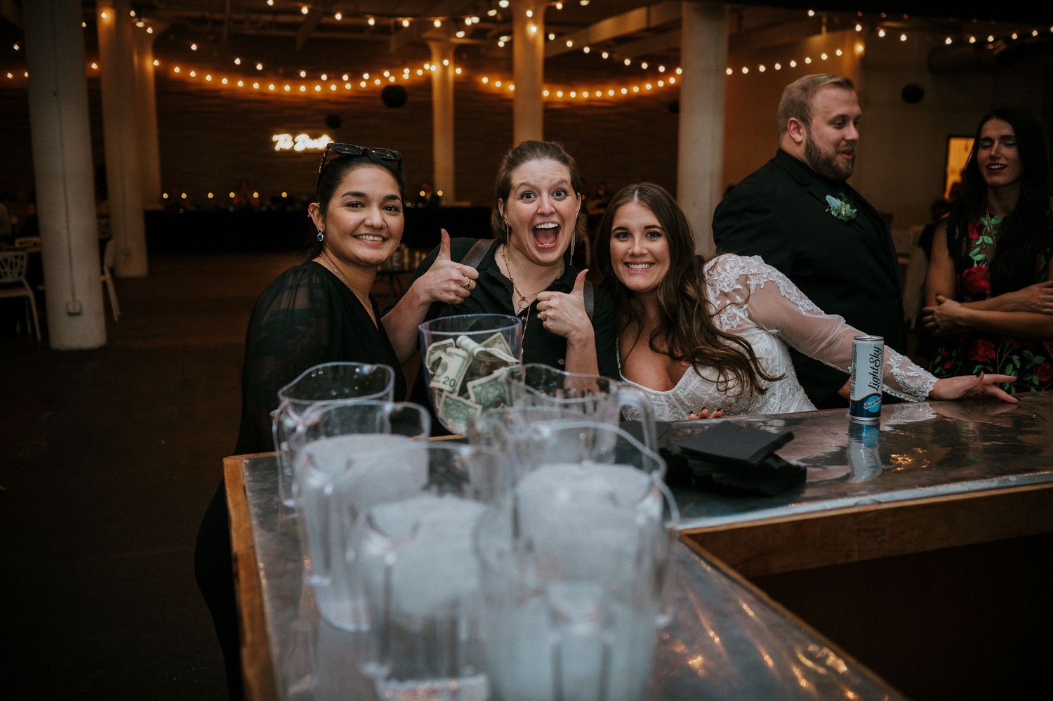 Three smiling women give thumbs up at a wedding reception bar with string lights glowing overhead.