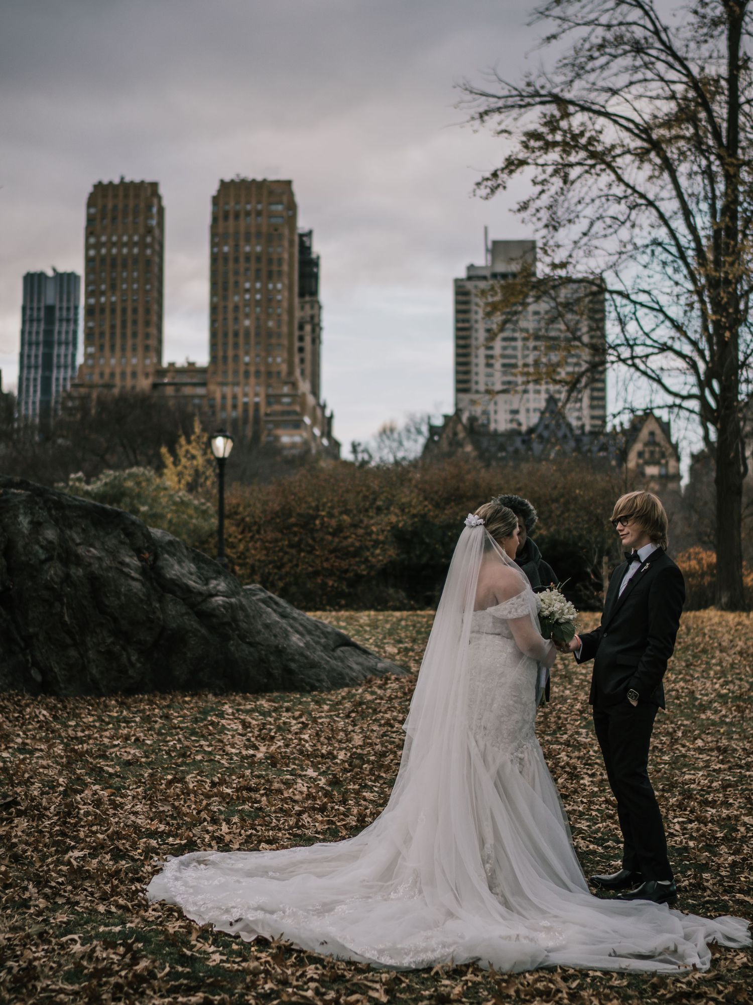 Bride and groom share a quiet moment amid golden autumn leaves with NYC skyline backdrop in Central Park.