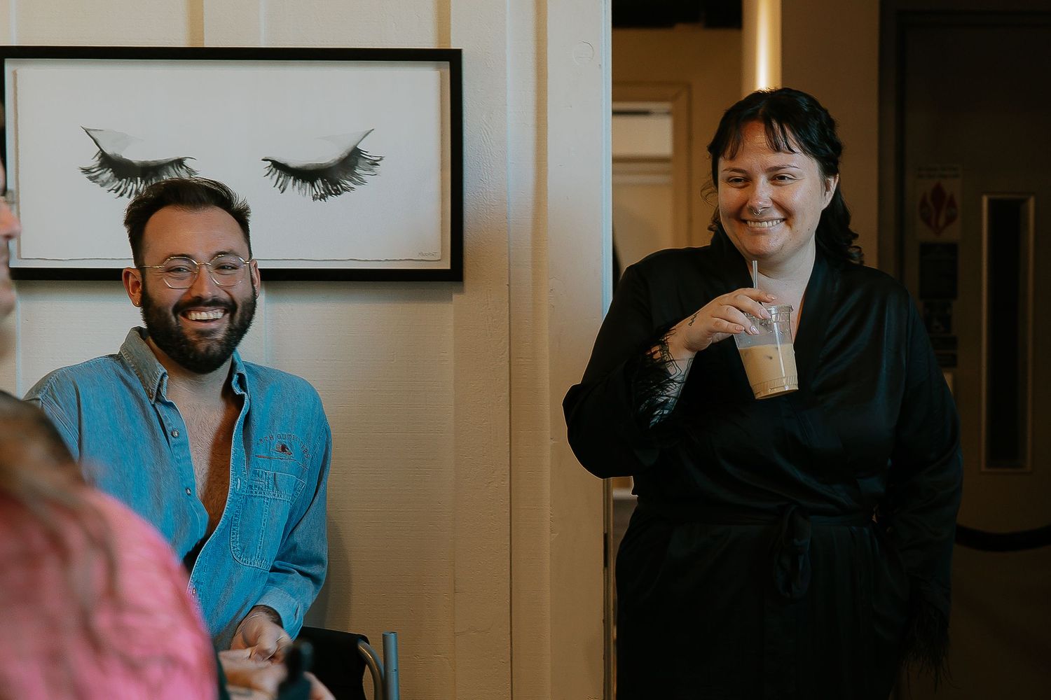 Two people laughing in a room with a decorative eyelash artwork on the wall.