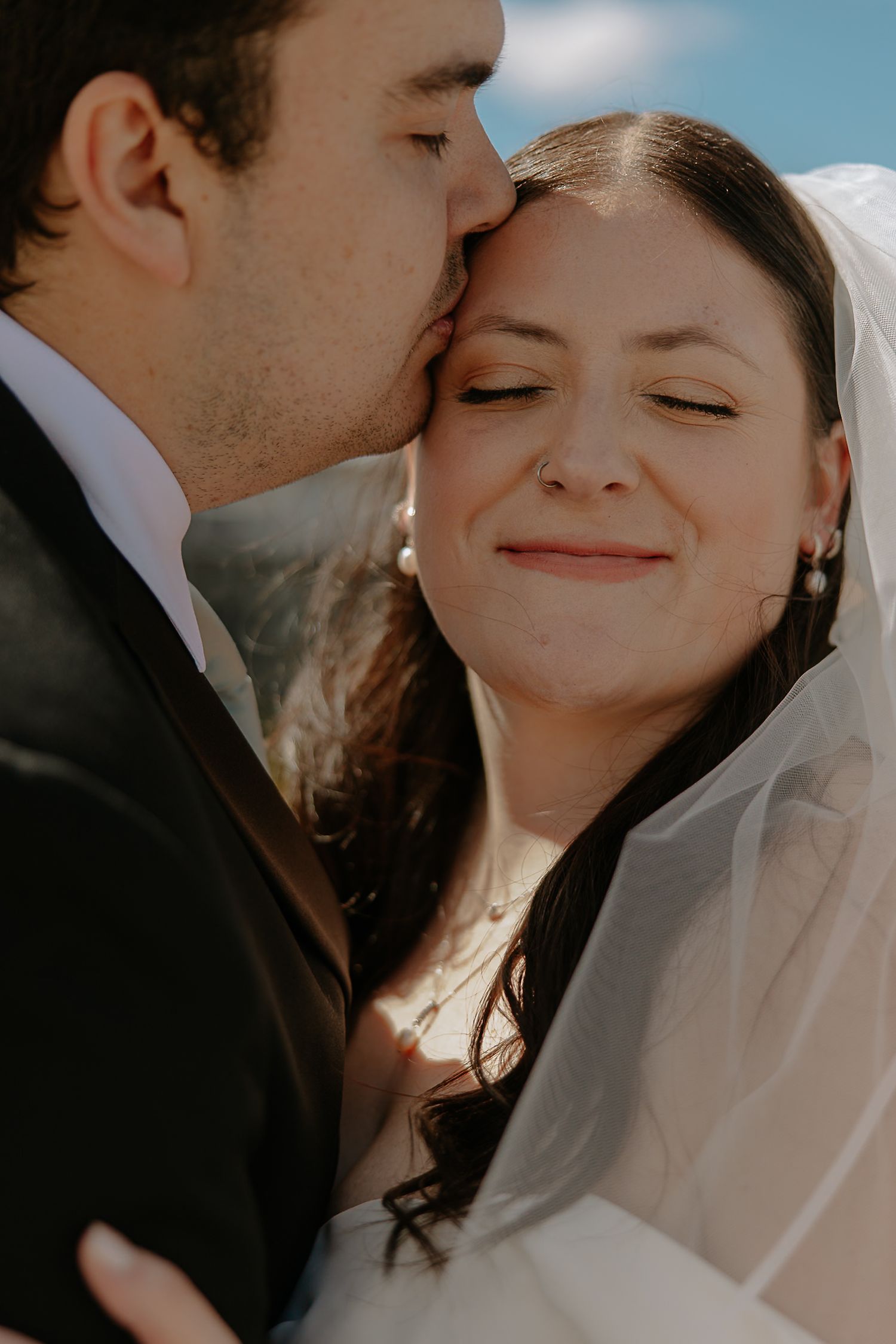 Groom tenderly kisses bride on the cheek as she smiles blissfully in her wedding veil outdoors.