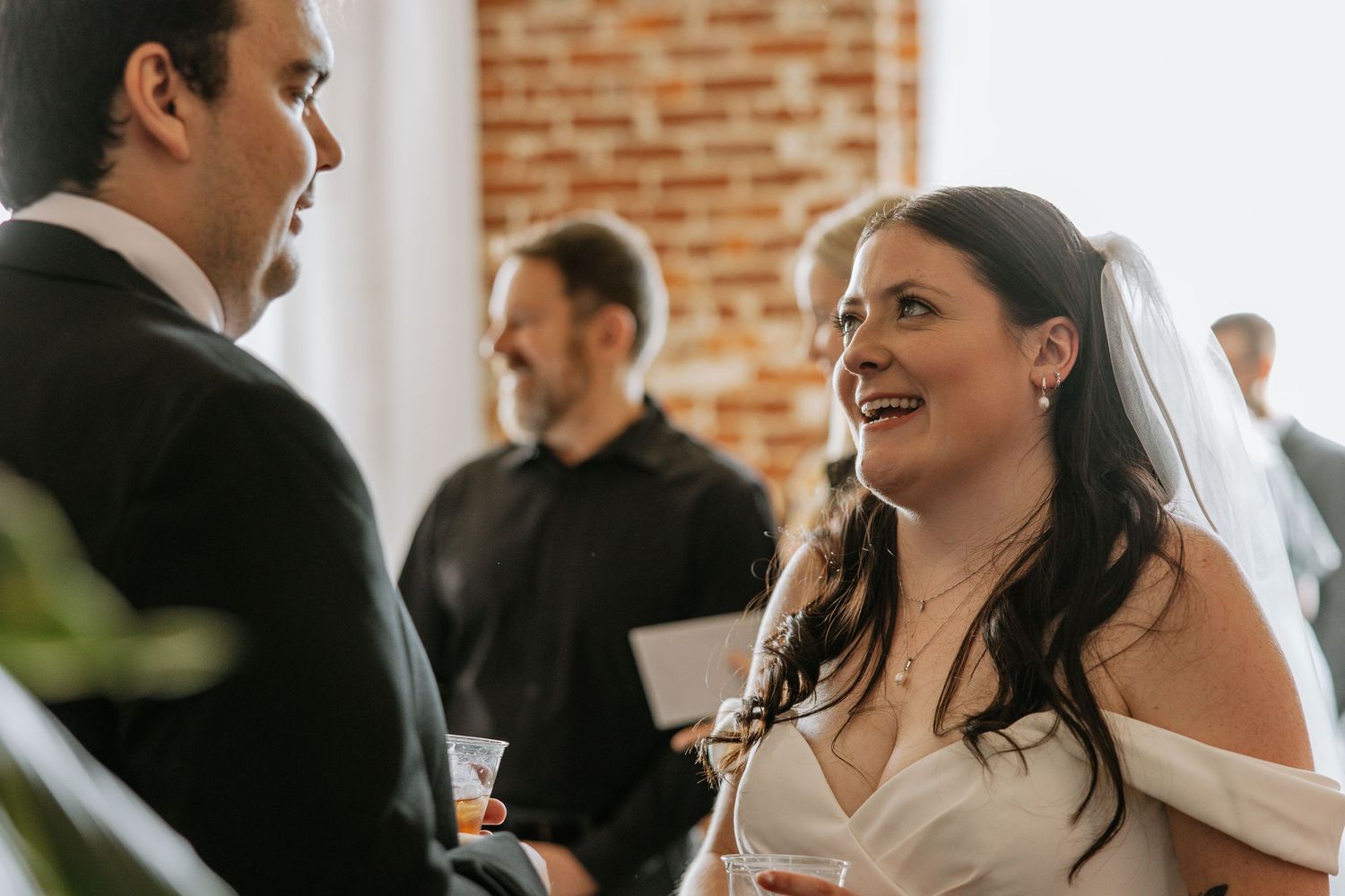 Bride in white off-shoulder gown smiles joyfully at groom during wedding reception with brick wall backdrop.
