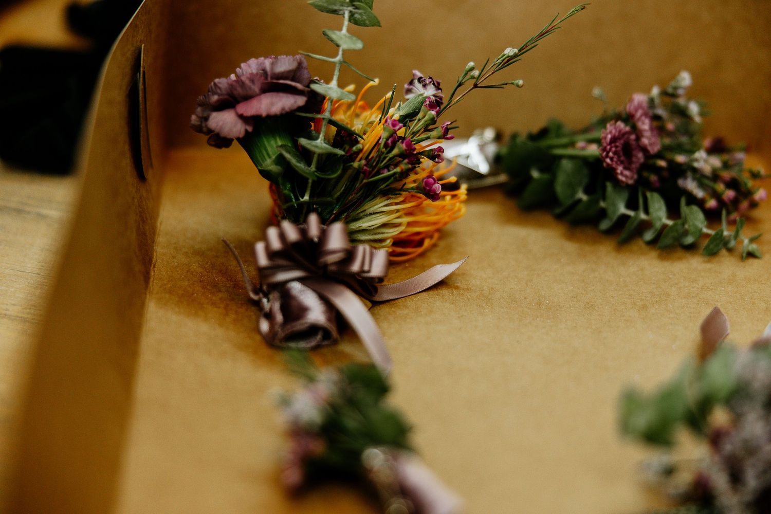 Elegant wedding boutonnieres and corsages with purple flowers and greenery arranged in a decorative box.