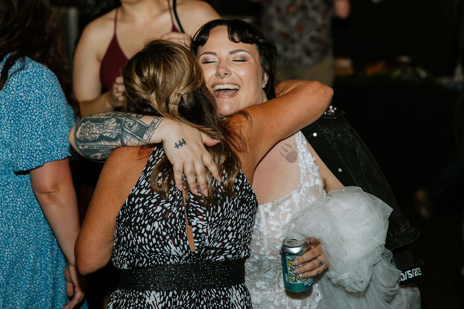Bride in white lace gown hugging guest in black patterned dress at wedding reception.
