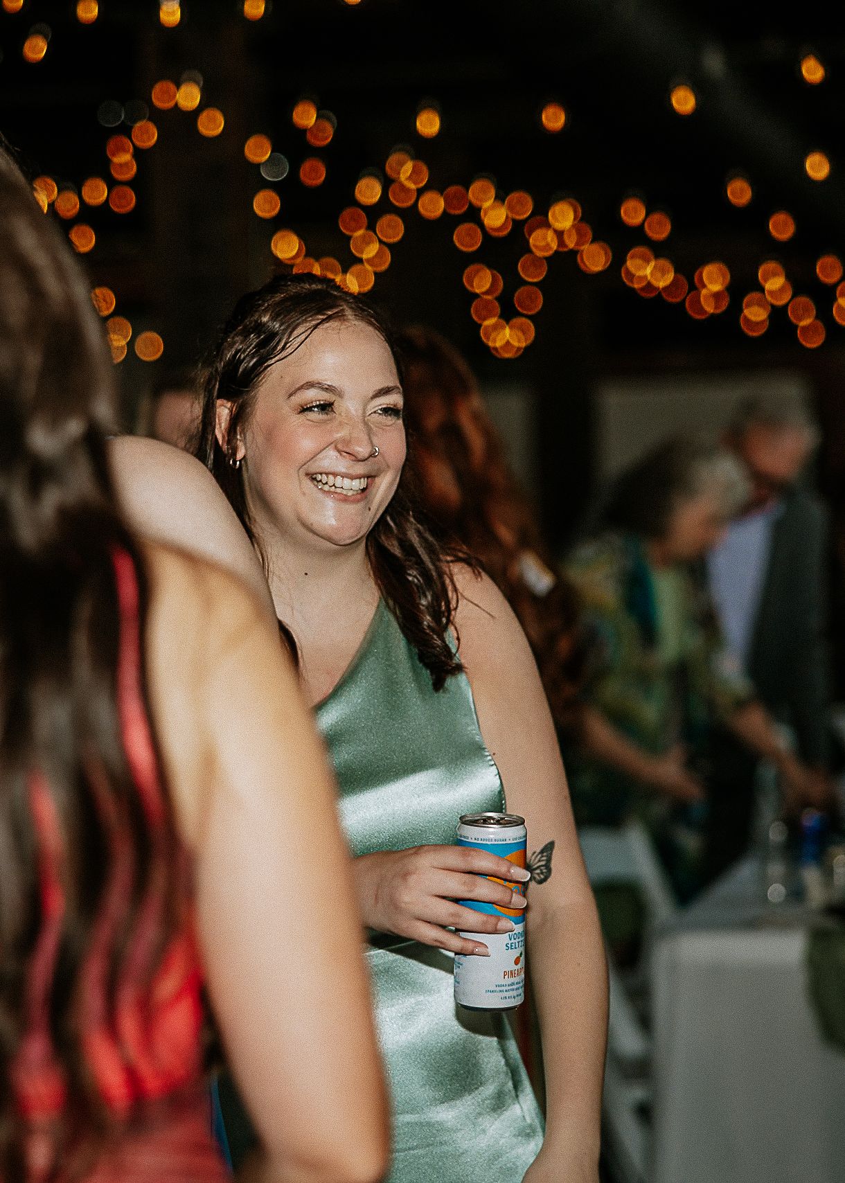 A smiling bridesmaid in a sage green satin dress holds a drink can at a wedding reception.