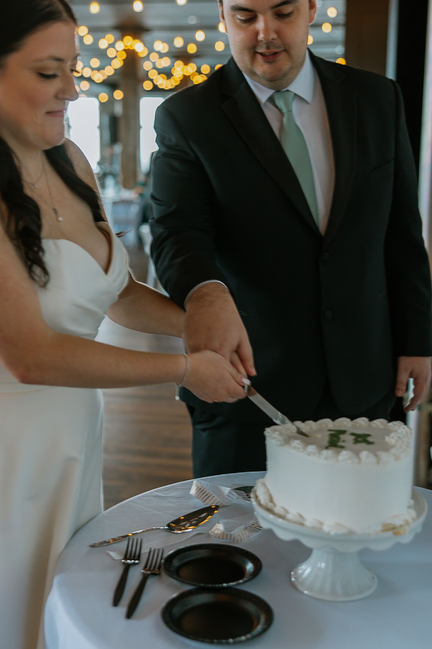 Bride and groom cut their white wedding cake together at the reception under string lights.
