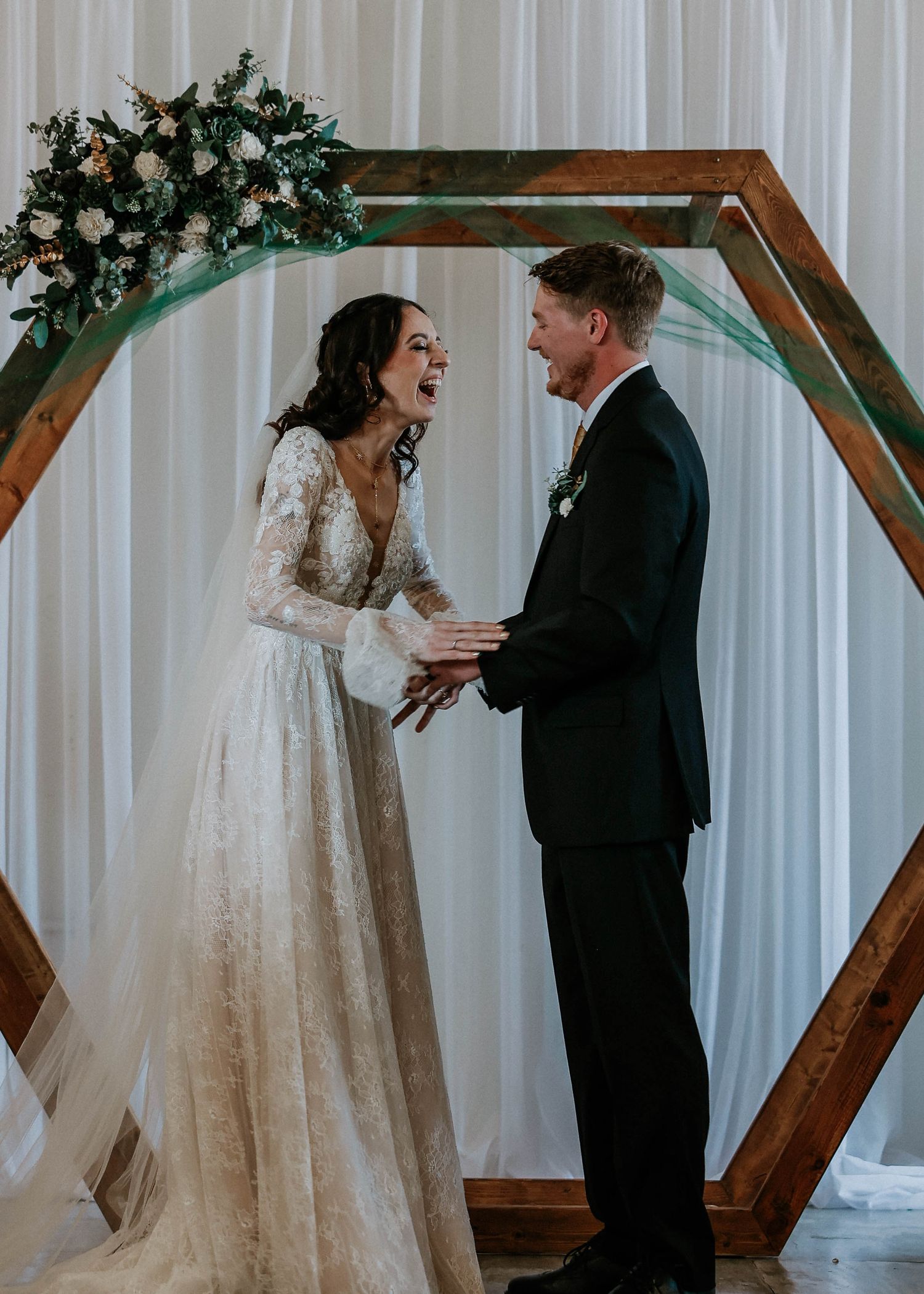 Bride and groom laughing joyfully at altar under hexagonal wooden arch with floral decor.