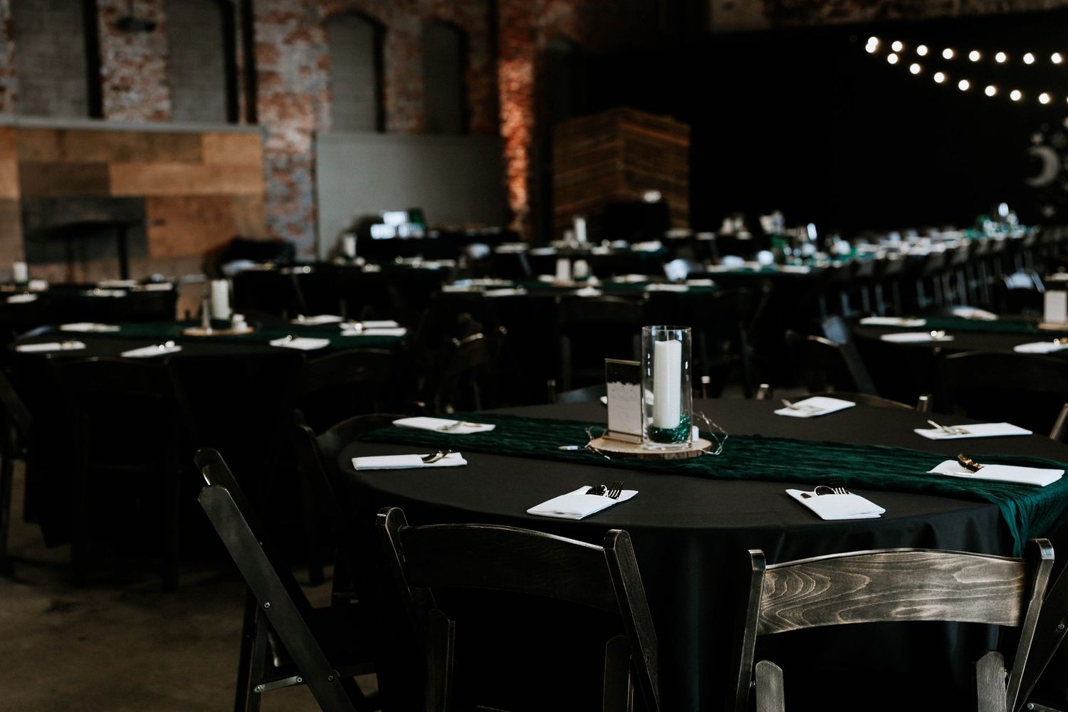 Elegant event venue with black round tables, green tablecloths, white candles, and string lights in a rustic brick setting.