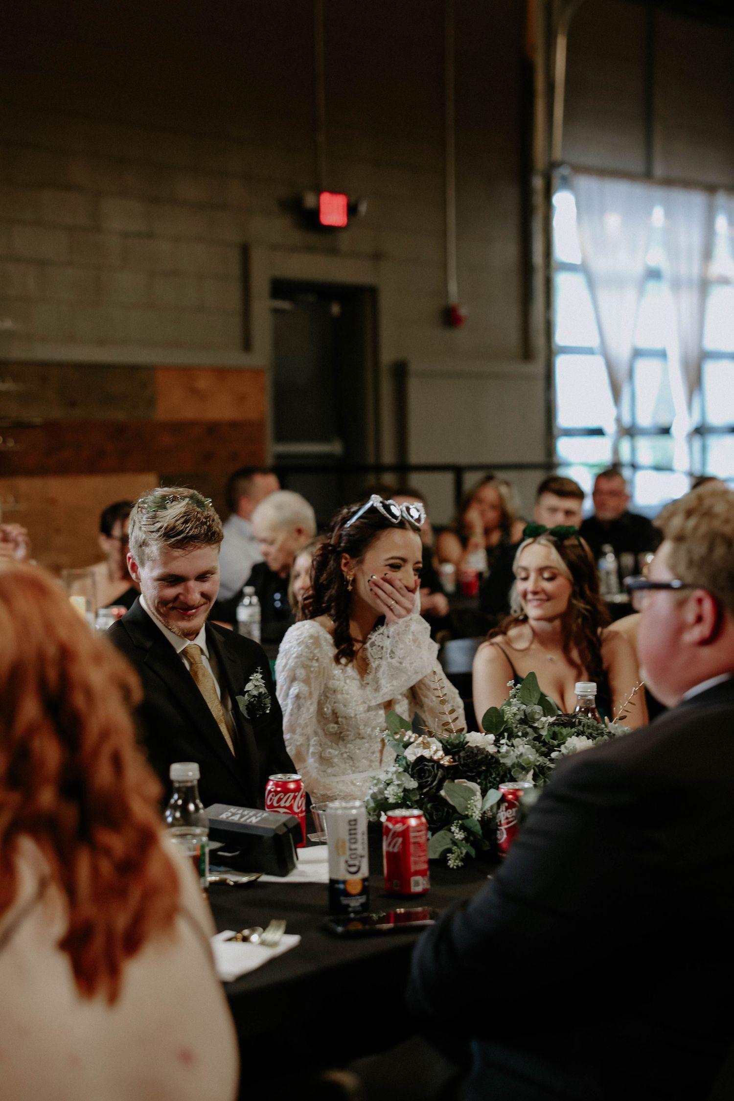 Bride laughing joyfully at wedding reception table with groom beside her, greenery centerpieces and guests in background.