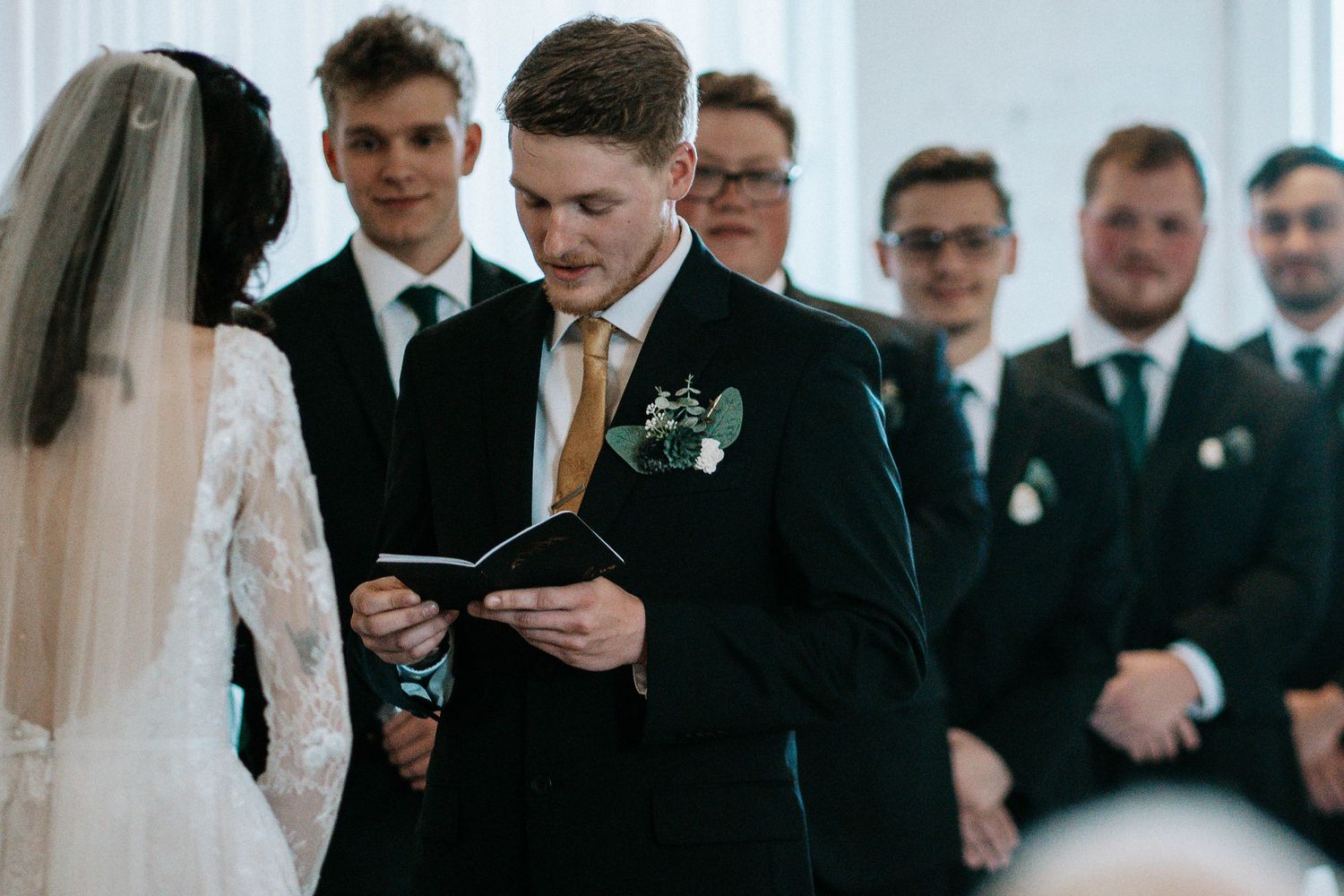 Groom reads wedding vows from a booklet at the altar, with groomsmen standing behind during the ceremony.