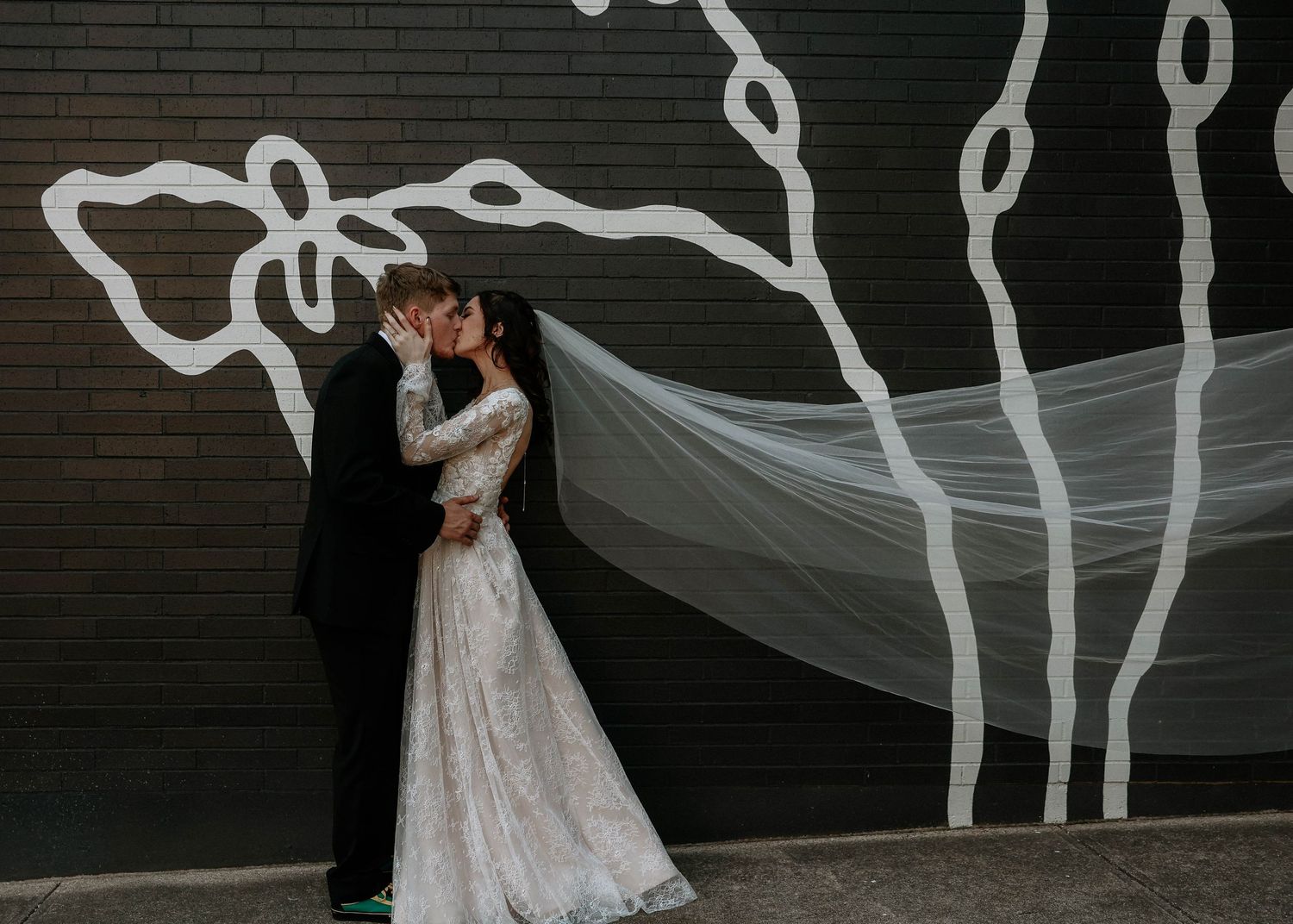 Bride and groom sharing a kiss in front of a dark brick wall with white floral mural, bride wearing lace gown with long veil.