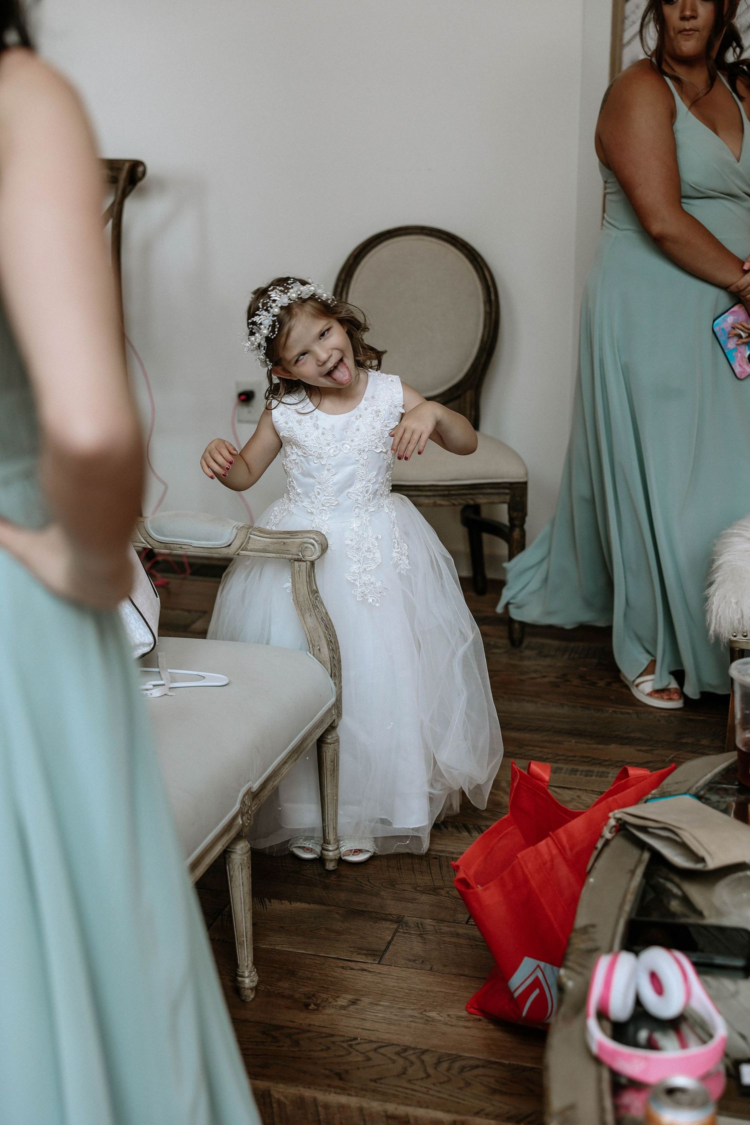 Adorable flower girl in white dress with floral crown laughing joyfully while seated at a wedding venue.